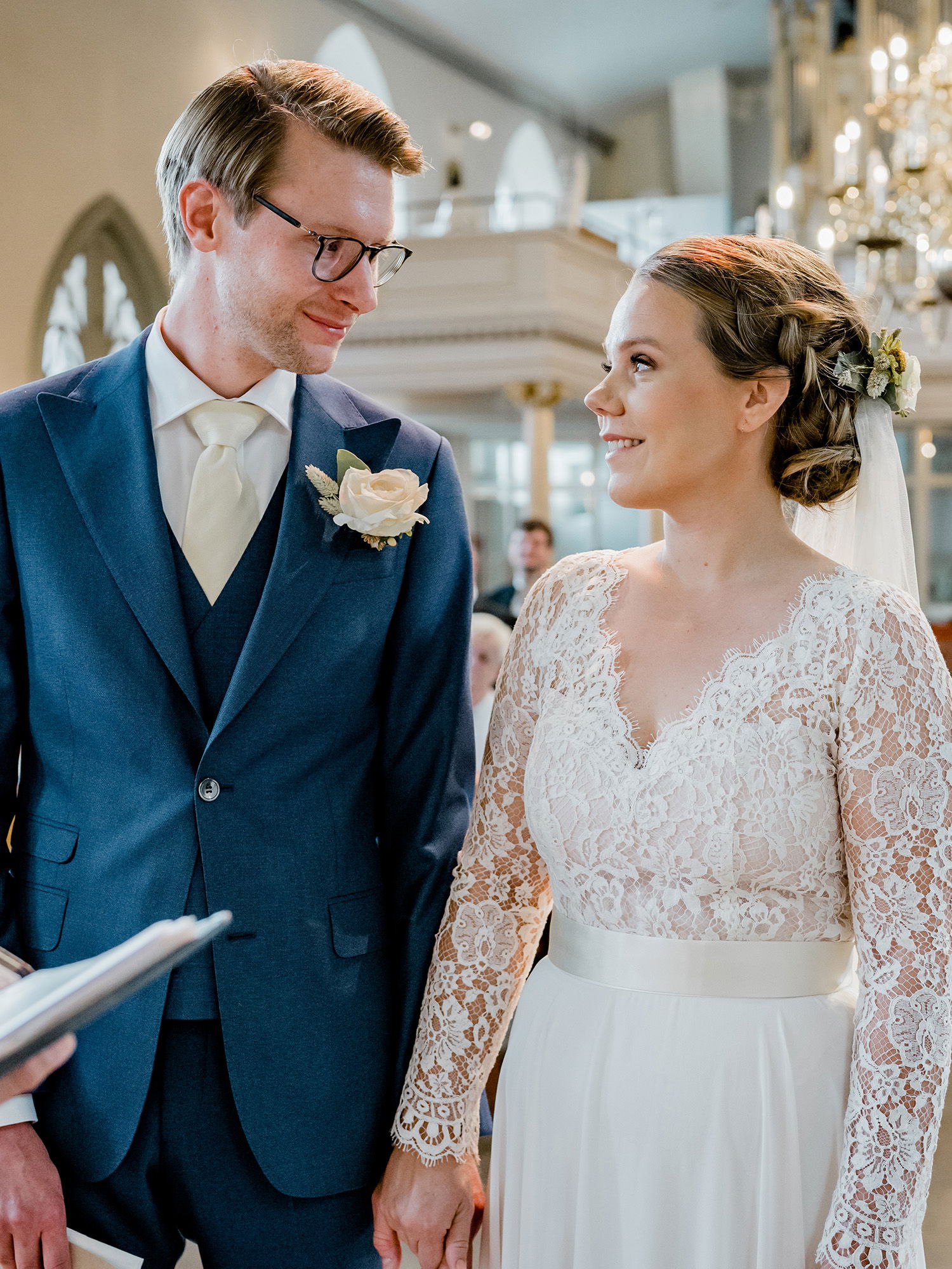 Das Brautpaar steht Hand in Hand während der kirchlichen Zeremonie. Die Braut in einem eleganten Spitzenkleid lächelt den Bräutigam an, der einen blauen Anzug trägt und sie liebevoll ansieht. Im Hintergrund leuchten die Fenster der Kirche.
