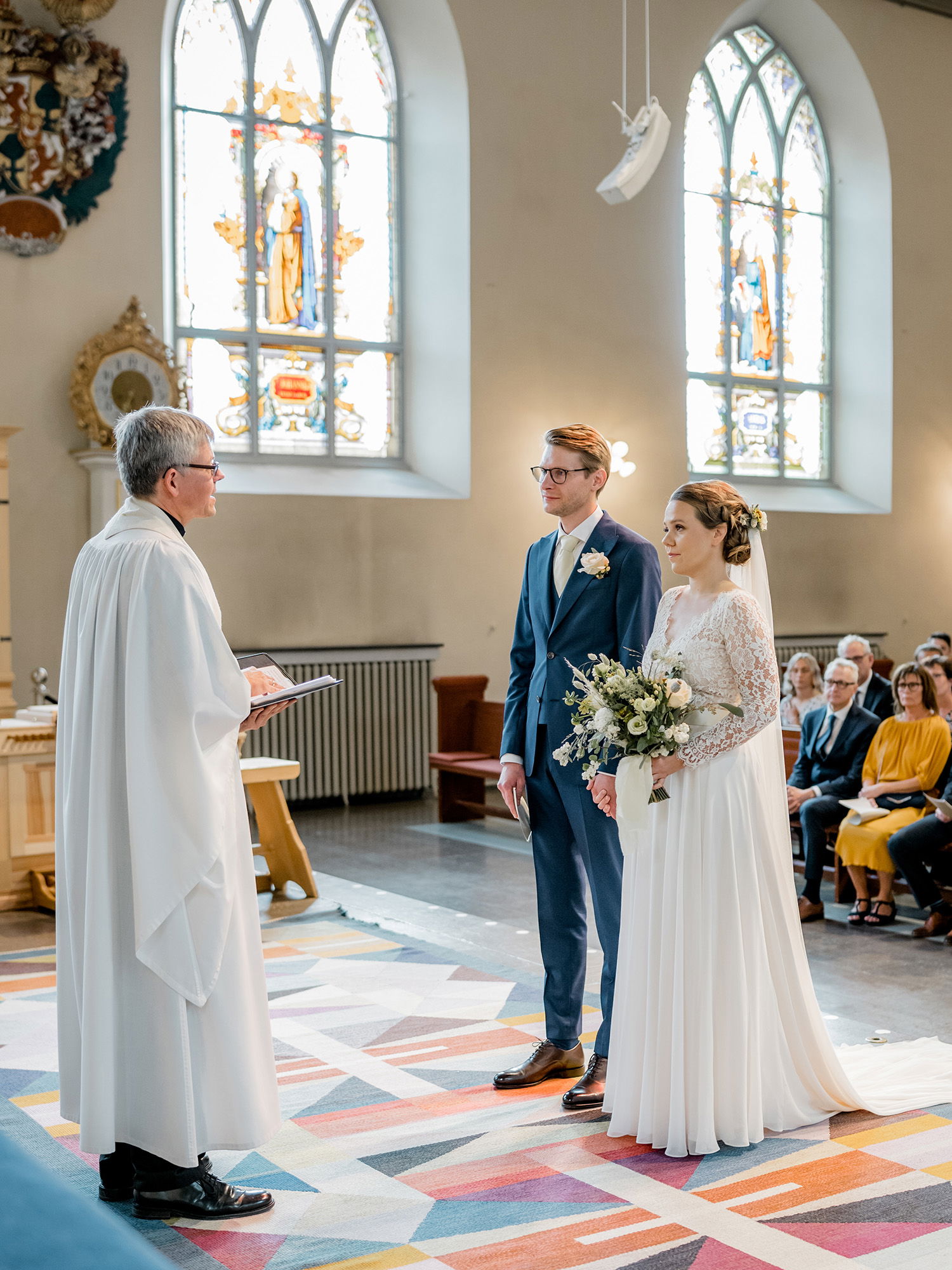Eine Braut und ein Bräutigam stehen in einer Kirche vor dem Priester und hören ihm während der Zeremonie zu. Die Braut trägt ein Kleid mit langen Spitzenärmeln und der Bräutigam einen eleganten blauen Anzug. Die Kulisse zeigt hohe Fenster mit bunten Glasmalereien.