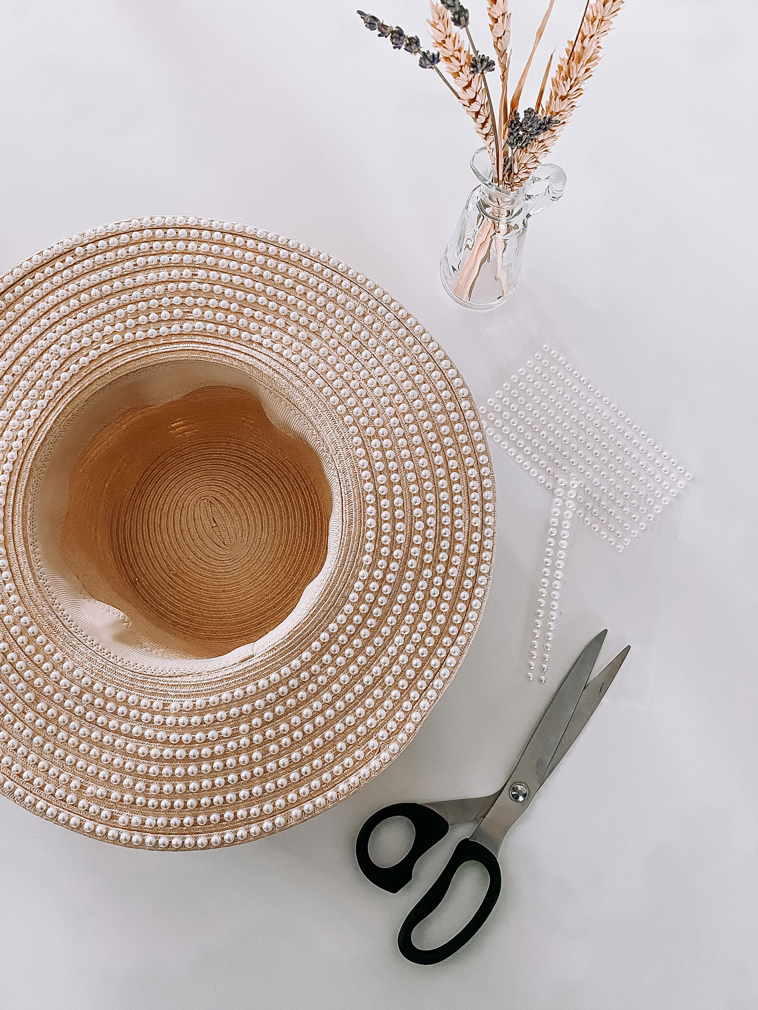 Ein DIY-Projekt mit einem Strohhut, der mit weißen Perlenstreifen verziert ist. Eine Schere und weitere Perlen auf einem Blatt sind als Bastelzubehör auf einer hellen Unterlage angeordnet.