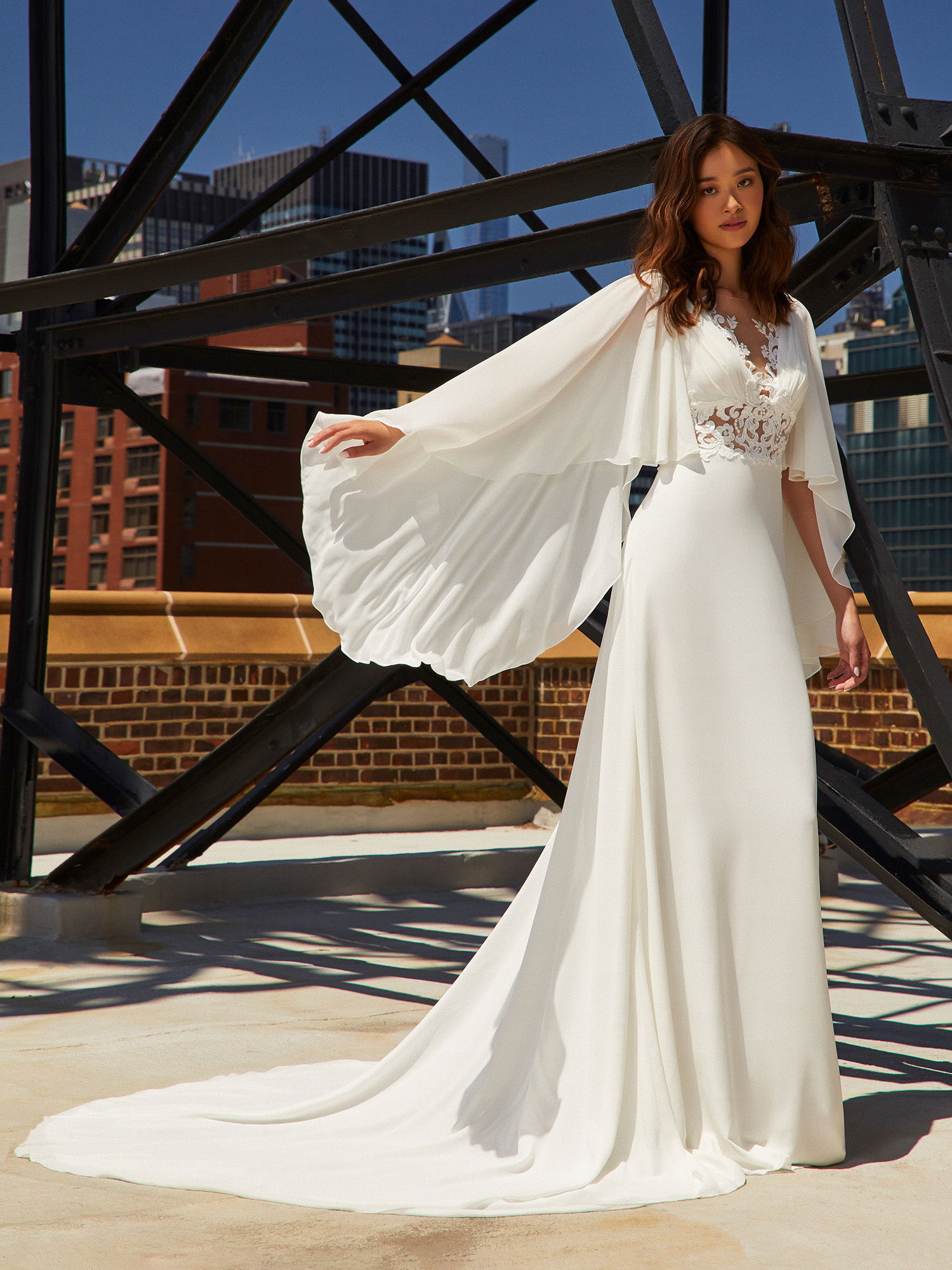 Die Braut posiert in einem eleganten Kleid mit bodenlanger Schleppe und weiten Flügelärmeln auf einer städtischen Terrasse. Ihr Outfit, kombiniert mit der architektonischen Kulisse und den Stahlstreben, schafft eine Mischung aus klassischer Eleganz und modernem Chic.