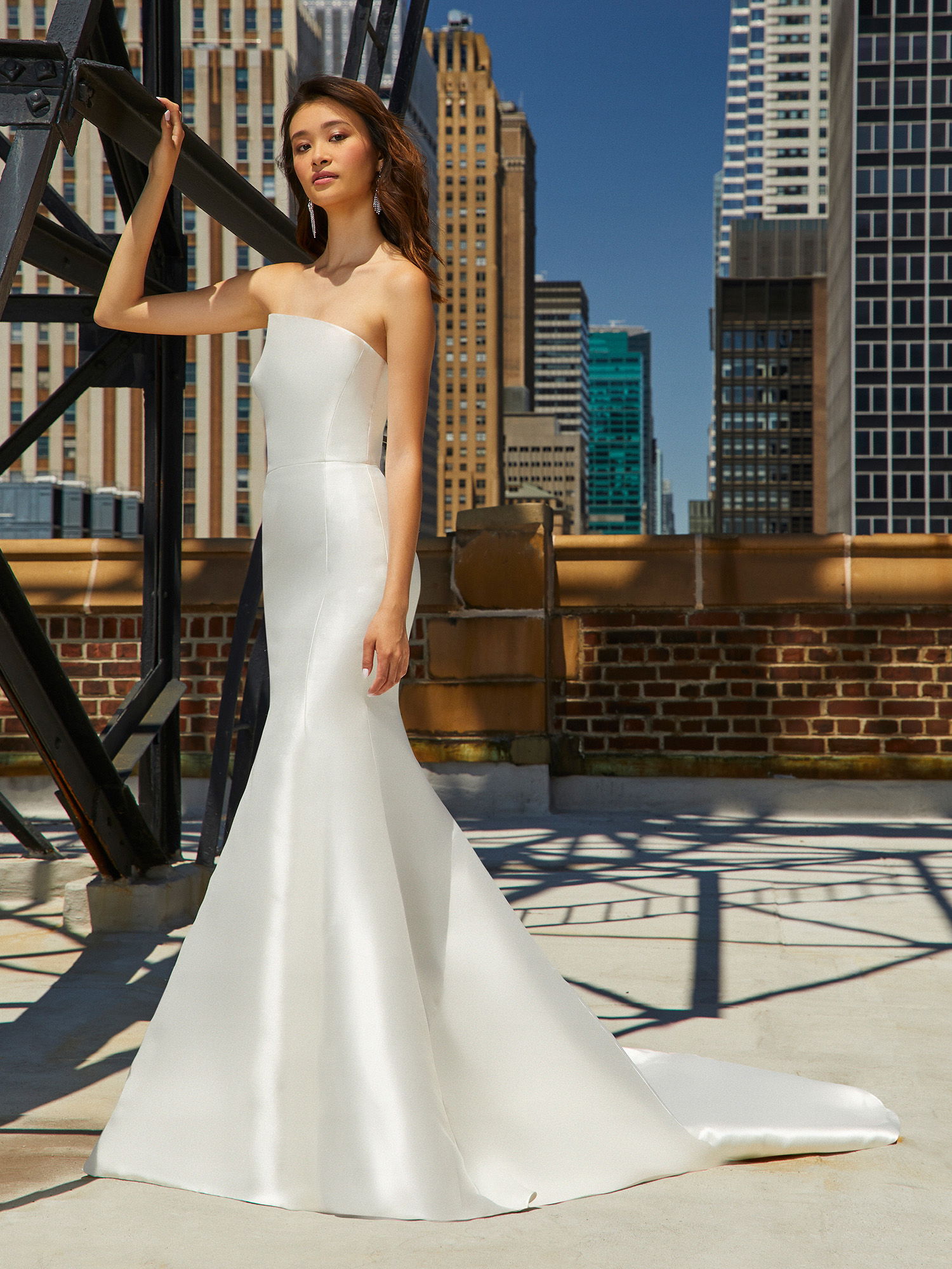 Eine Frau in einem minimalistischen, trägerlosen Brautkleid steht auf einer Terrasse im Freien, umgeben von Wolkenkratzern. Ihr Kleid fällt in einer fließenden Linie bis zum Boden, und die stahlgraue Kulisse der Stadt verstärkt den modernen, schicken Stil der Aufnahme.