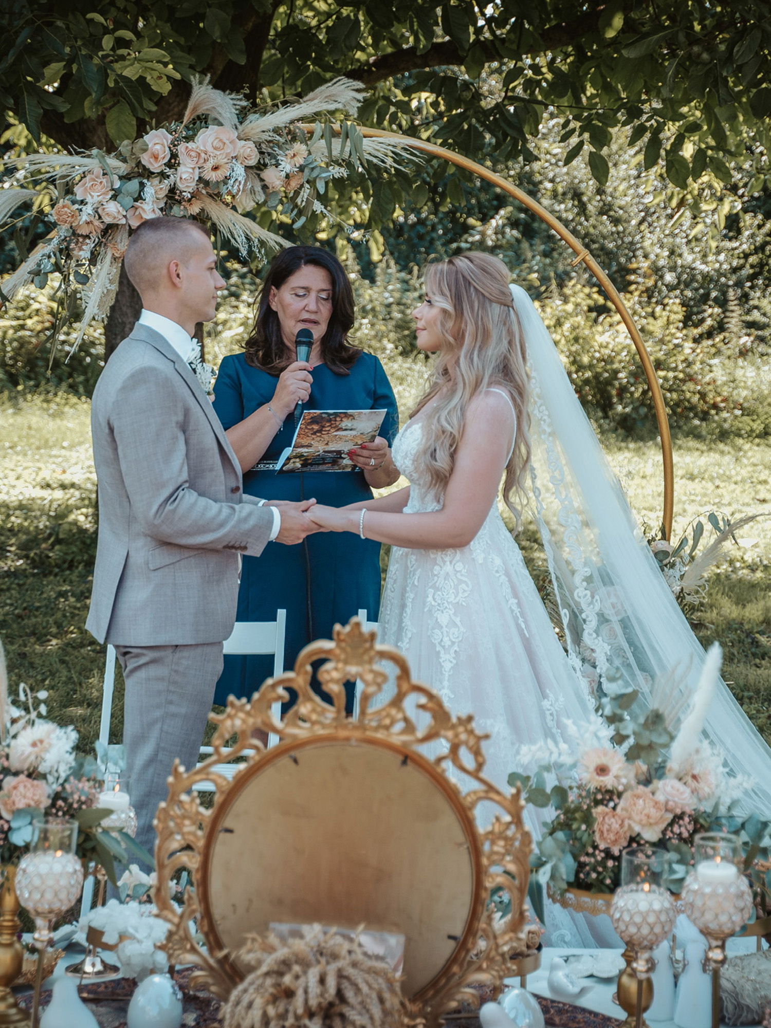 Eine Hochzeitszeremonie im Freien mit einem Brautpaar, das sich unter einem runden Blumenbogen die Hände hält. Die Traurednerin in Blau liest aus einem Buch vor. Vor dem Paar befindet sich ein festlich geschmückter Tisch mit Blumen und goldenen Elementen.