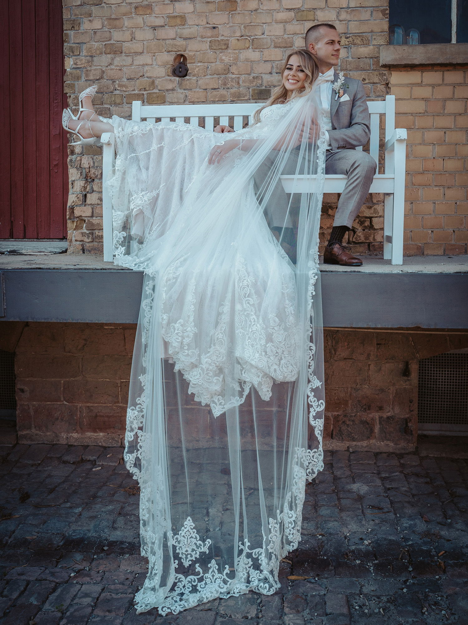 Das Hochzeitspaar sitzt zusammen auf einer weißen Bank vor einer rustikalen Backsteinfassade. Die Braut lächelt und streckt ihre Beine aus, während ihr langer Schleier über die Bank hängt. Der Bräutigam trägt einen grauen Anzug und schaut entspannt zur Seite.