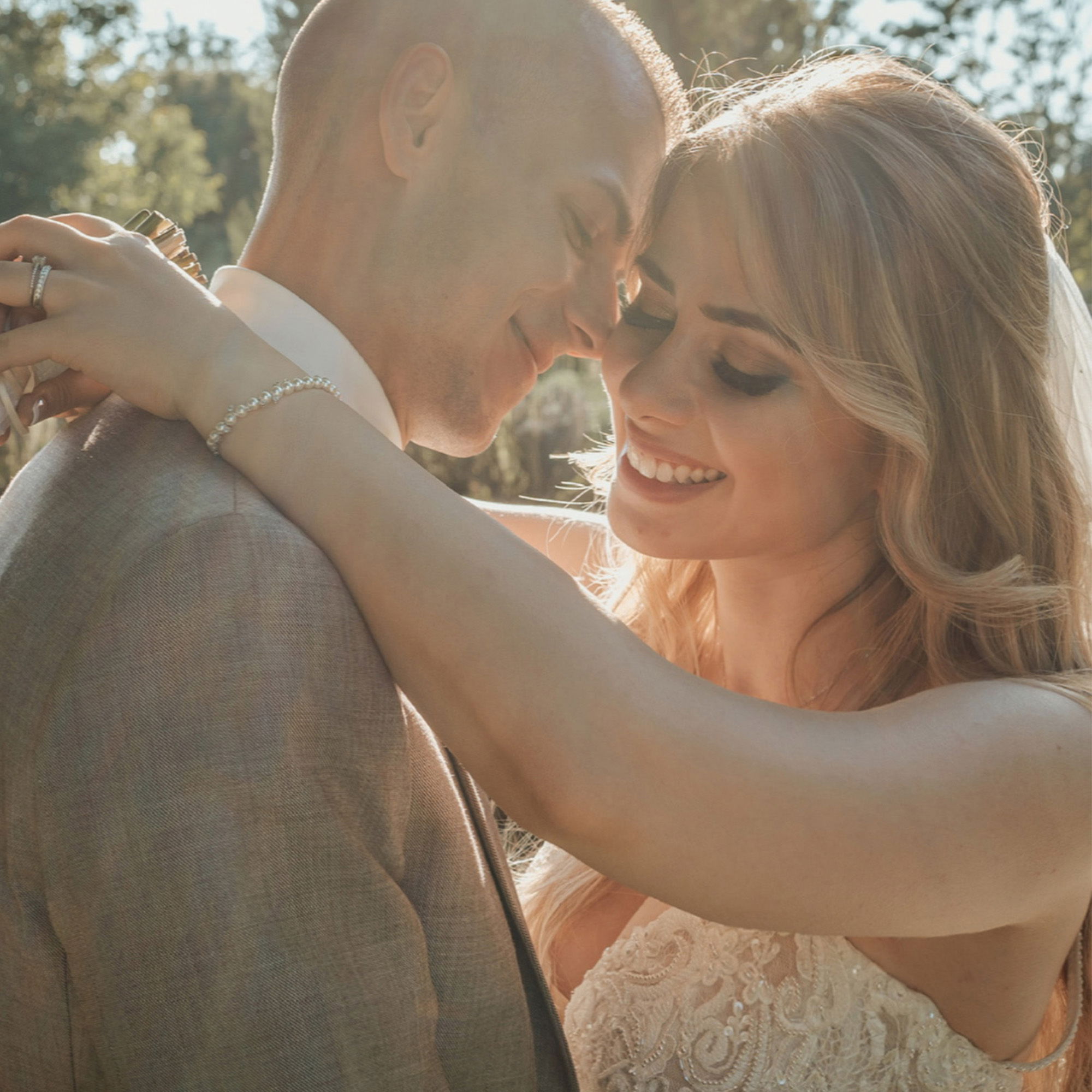 Ein verliebtes Hochzeitspaar umarmt sich und lächelt einander zu. Die Braut in einem weißen Hochzeitskleid legt ihre Arme um den Bräutigam, der einen eleganten Anzug trägt. Sie befinden sich in einem sonnigen Garten.