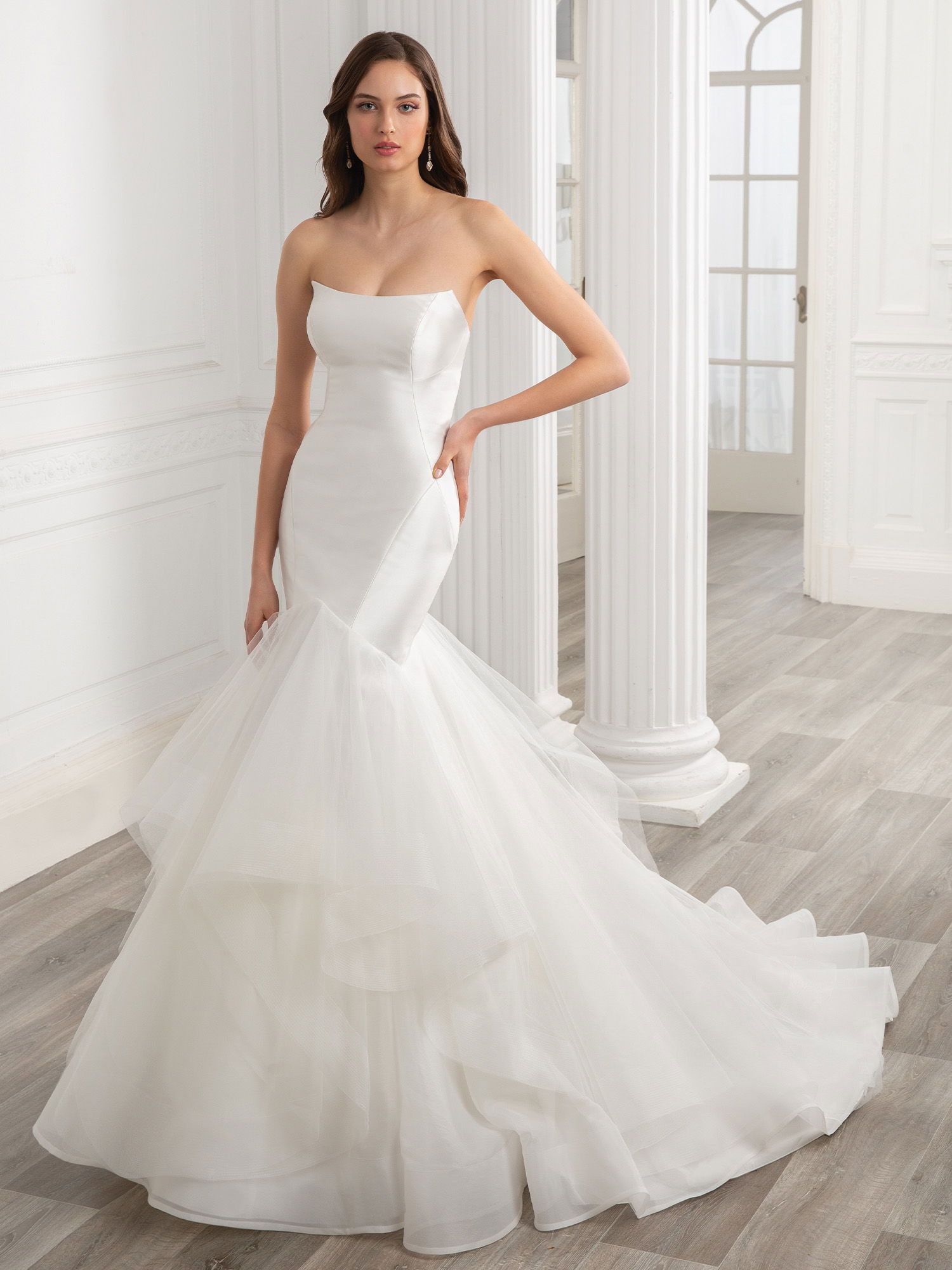 Eine Braut in einem modernen Meerjungfrauen-Brautkleid aus Seide und Tüll posiert selbstbewusst vor weißen Säulen in einem hellen Raum. Ihr Kleid betont ihre schlanke Silhouette, während der voluminöse Rock sanft fällt.