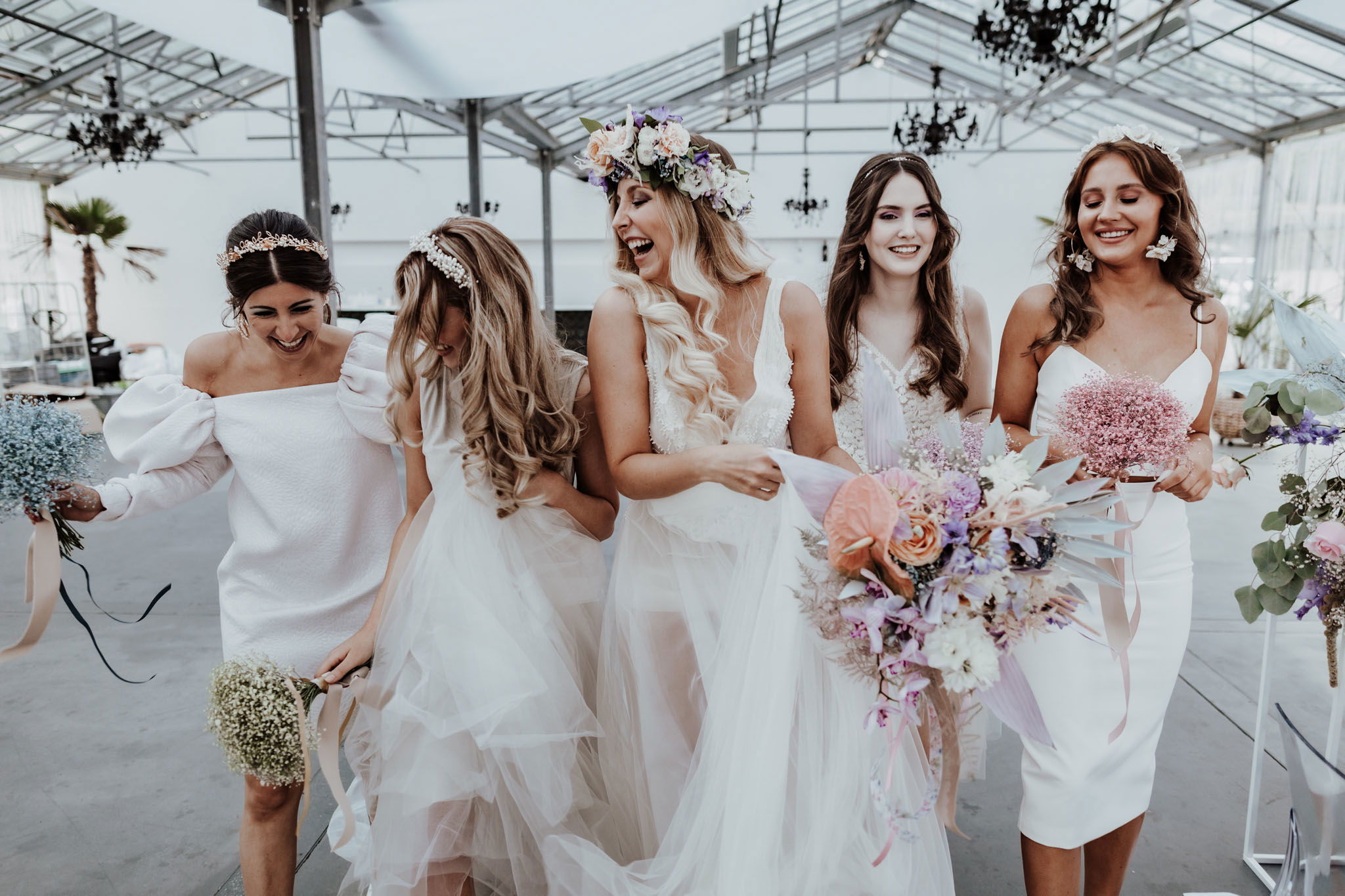 Fünf Bräute und Brautjungfern in stilvollen weißen Hochzeitskleidern, die in einer hellen, eleganten Halle fröhlich gemeinsam den Moment genießen. Blumige Accessoires und lockere Haare verleihen ihnen einen natürlichen Look.