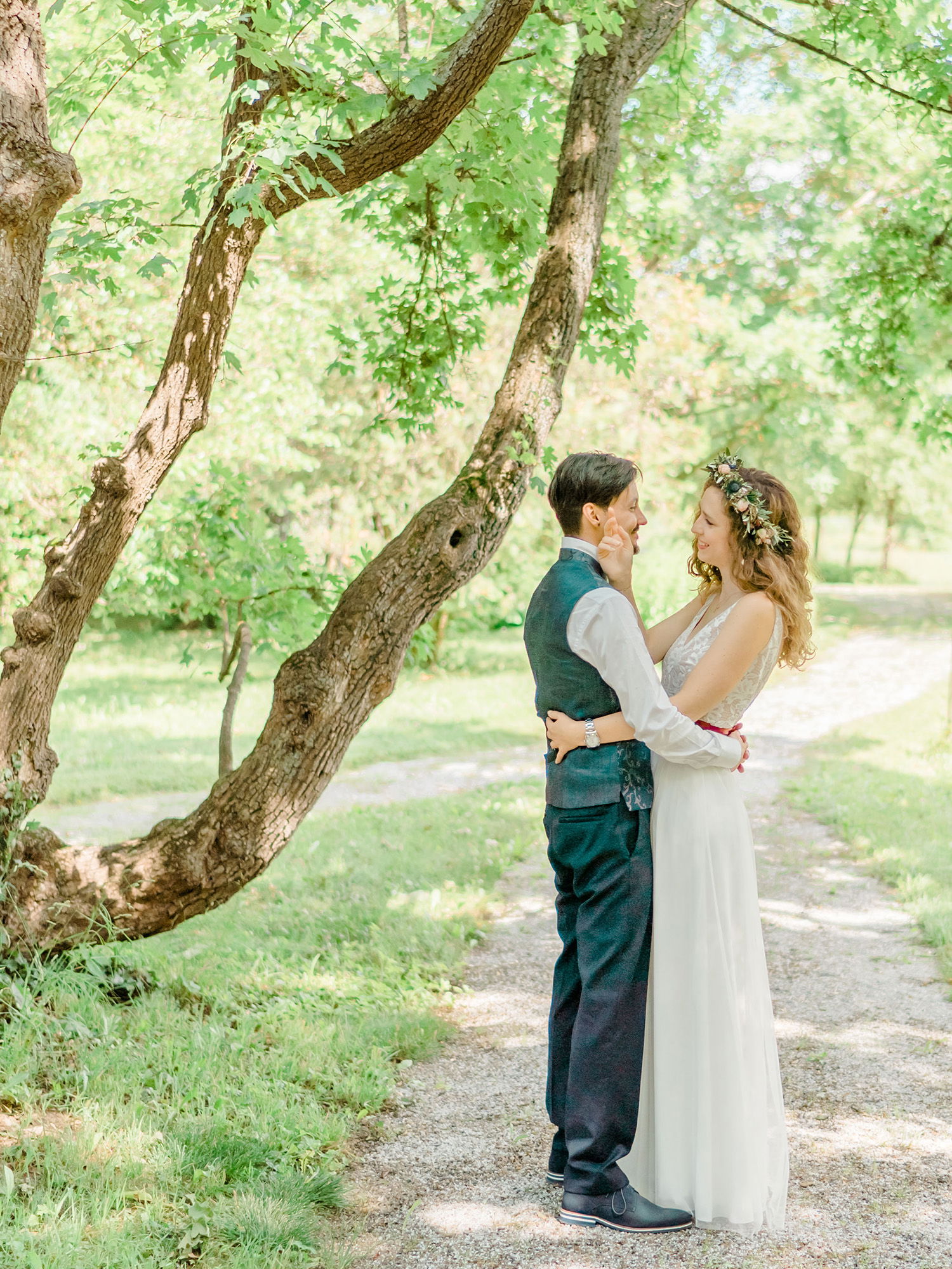 Das Brautpaar steht inmitten einer malerischen Naturkulisse mit üppigem Laub. Die Braut berührt sanft das Gesicht des Bräutigams, während sie sich innig in die Augen schauen. Beide wirken in ihrem natürlichen Hochzeitslook entspannt und glücklich.