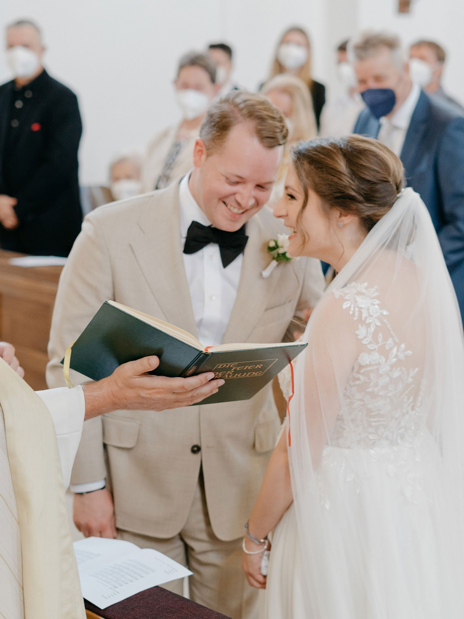 Der Bräutigam und die Braut lächeln sich während der Trauung an, während der Priester eine Bibel in der Hand hält. Die Braut trägt einen langen Schleier und ein Spitzenkleid. Die Zeremonie findet in einer Kirche mit maskierten Gästen statt.