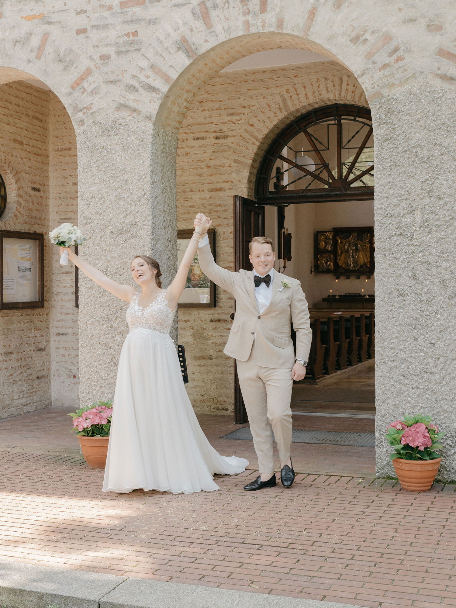 Ein Brautpaar jubelt nach der Trauung vor einer alten Kirche. Die Braut hebt ihren Blumenstrauß in die Luft, während der Bräutigam in einem hellen Anzug neben ihr steht. Beide lächeln, umgeben von antiker Architektur und Blumendekoration.