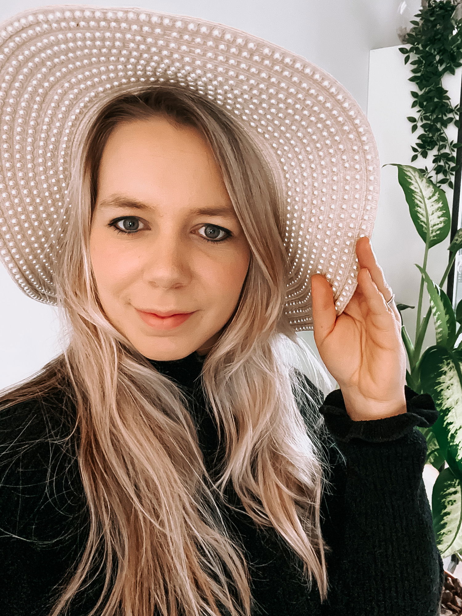 Eine Frau mit welligem, hellblondem Haar trägt einen perlenbesetzten Hut und lächelt leicht in die Kamera. Der Perlenhut bildet einen markanten Kontrast zu ihrem schwarzen Pullover.