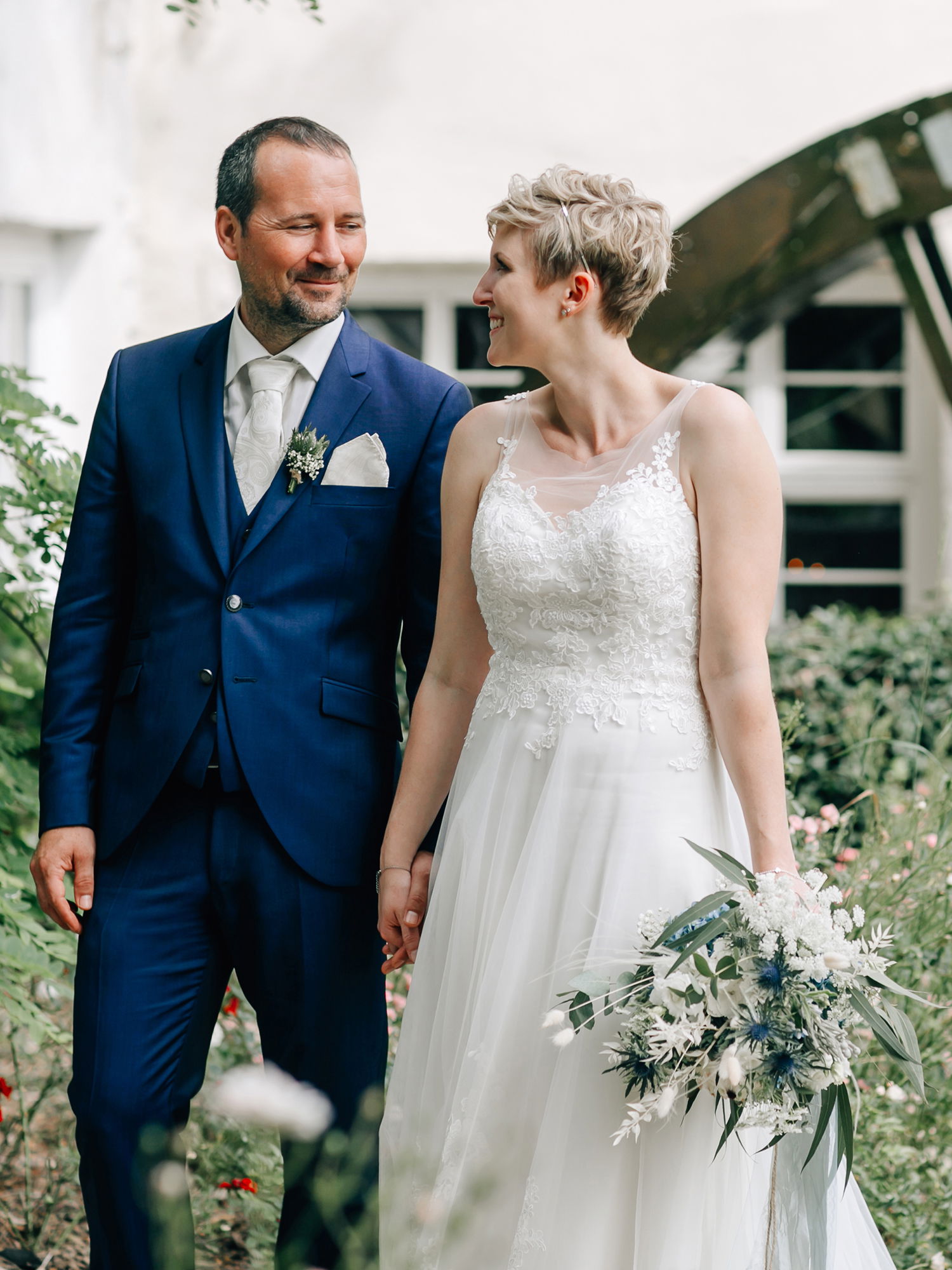 Die Braut in einem ärmellosen weißen Spitzenkleid und der Bräutigam in einem stilvollen blauen Anzug gehen gemeinsam durch einen üppigen Garten. Die Braut trägt einen Blumenstrauß mit weißen und grünen Akzenten. Sie sehen sich an, während sie ihre Hände halten und einander zulächeln.