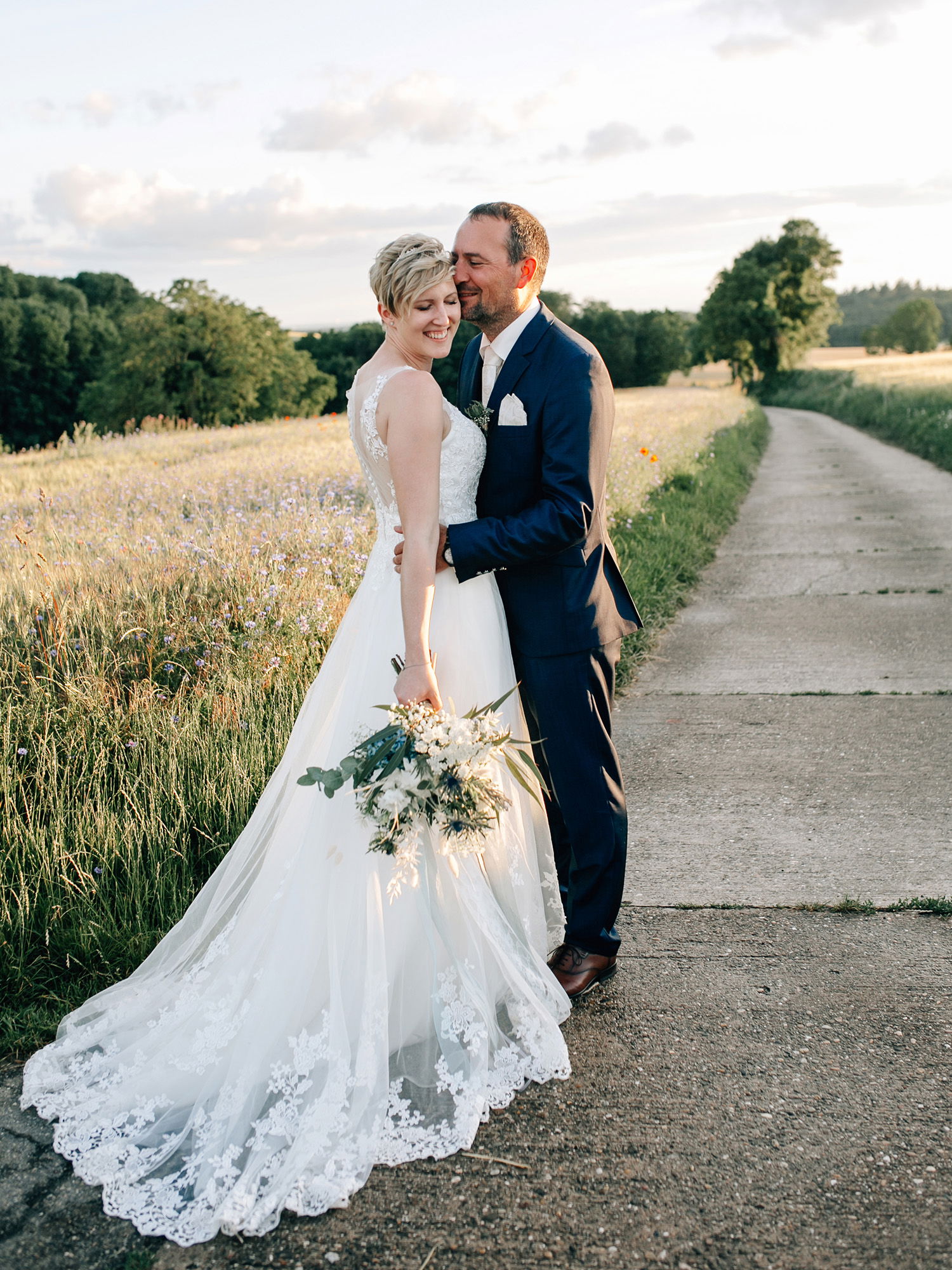 Ein Brautpaar posiert in einer malerischen Wiesenlandschaft bei Sonnenuntergang. Die Braut im weißen Kleid lächelt glücklich, während der Bräutigam im eleganten Anzug sie sanft umarmt. Sie hält einen Blumenstrauß mit weißen und grünen Akzenten in der Hand.