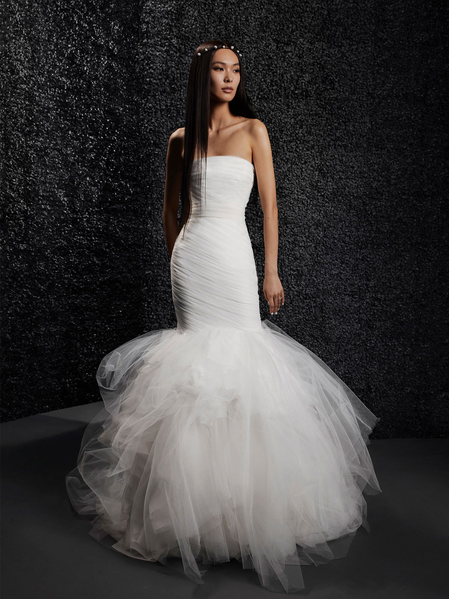 Die Braut posiert in einem stilvollen trägerlosen Kleid, das mit zarten Falten am Oberteil und einem dramatisch voluminösen Tüllrock im Meerjungfrauenstil verziert ist. Ihre langen, glatten Haare sind mit einem dezenten Perlenband geschmückt.