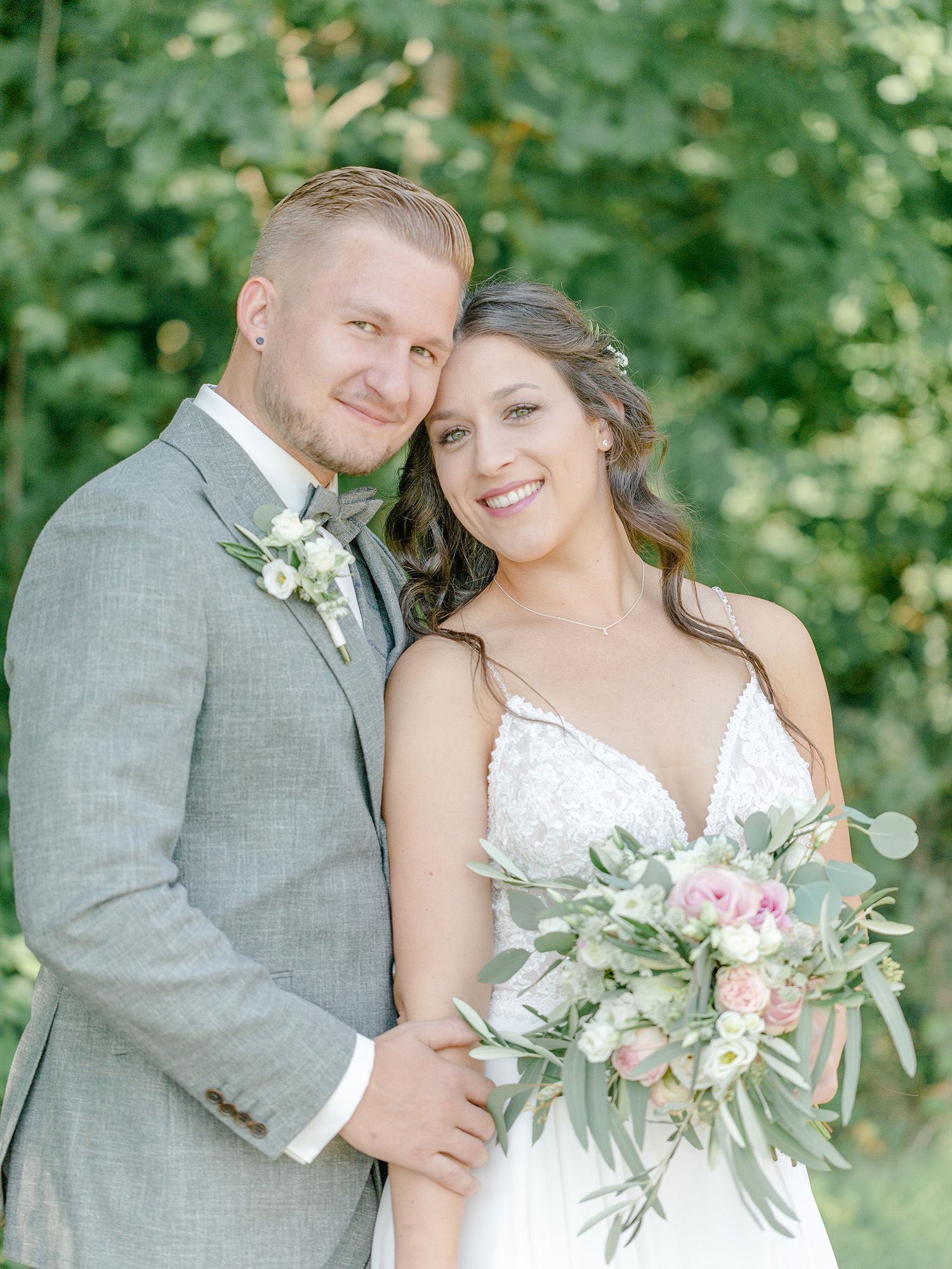 Nahaufnahme eines frisch verheirateten Paares, das sich in einem Waldstück aneinander lehnt. Der Bräutigam hat einen grauen Anzug an, während die Braut ein florales Bouquet mit rosa Akzenten hält und in einem eleganten Kleid strahlt.