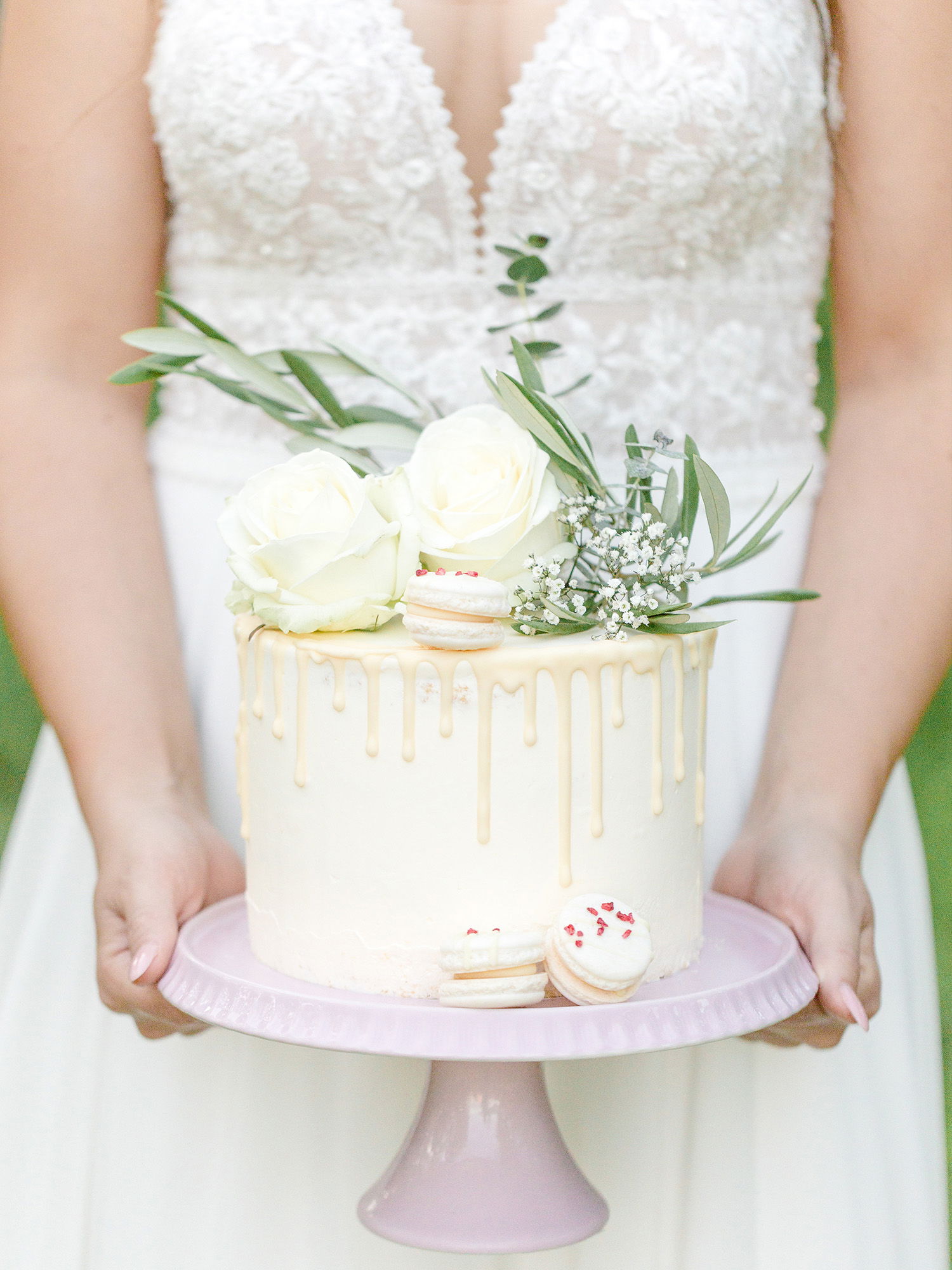 Nahaufnahme einer Hochzeitstorte auf einer rosa Tortenplatte, gehalten von einer Braut in einem Spitzenkleid. Die Torte ist mit weißen Blumen, Macarons und frischen Blättern dekoriert, während die glasige Glasur elegant herunterläuft.