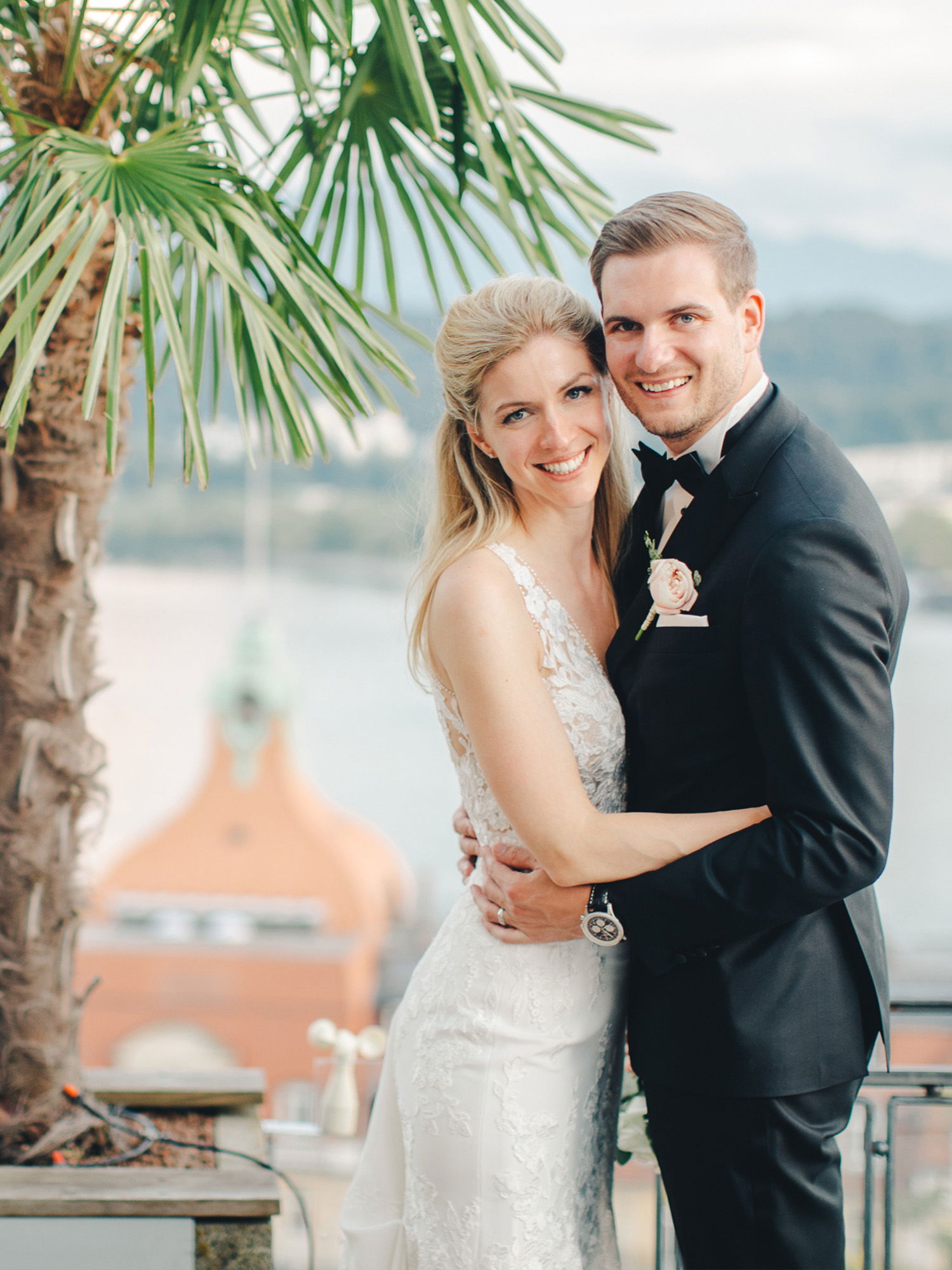 Ein Hochzeitspaar posiert vor einer malerischen Kulisse mit Palmen. Die Braut, in einem weißen Kleid mit Spitzen, wird vom Bräutigam in einem schwarzen Anzug fest umarmt. Beide strahlen und stehen vor einem Aussichtspunkt mit Blick auf ein historisches Gebäude.