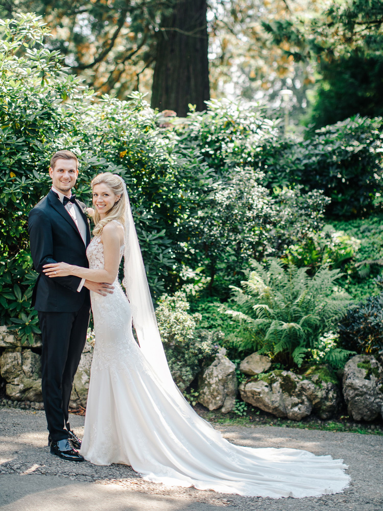 Ein Hochzeitspaar posiert inmitten eines grünen Gartens. Die Braut in einem schlichten Hochzeitskleid mit Schleppe lehnt sich an den Bräutigam, der einen klassischen schwarzen Anzug trägt. Im Hintergrund sind Bäume und Sträucher zu sehen.