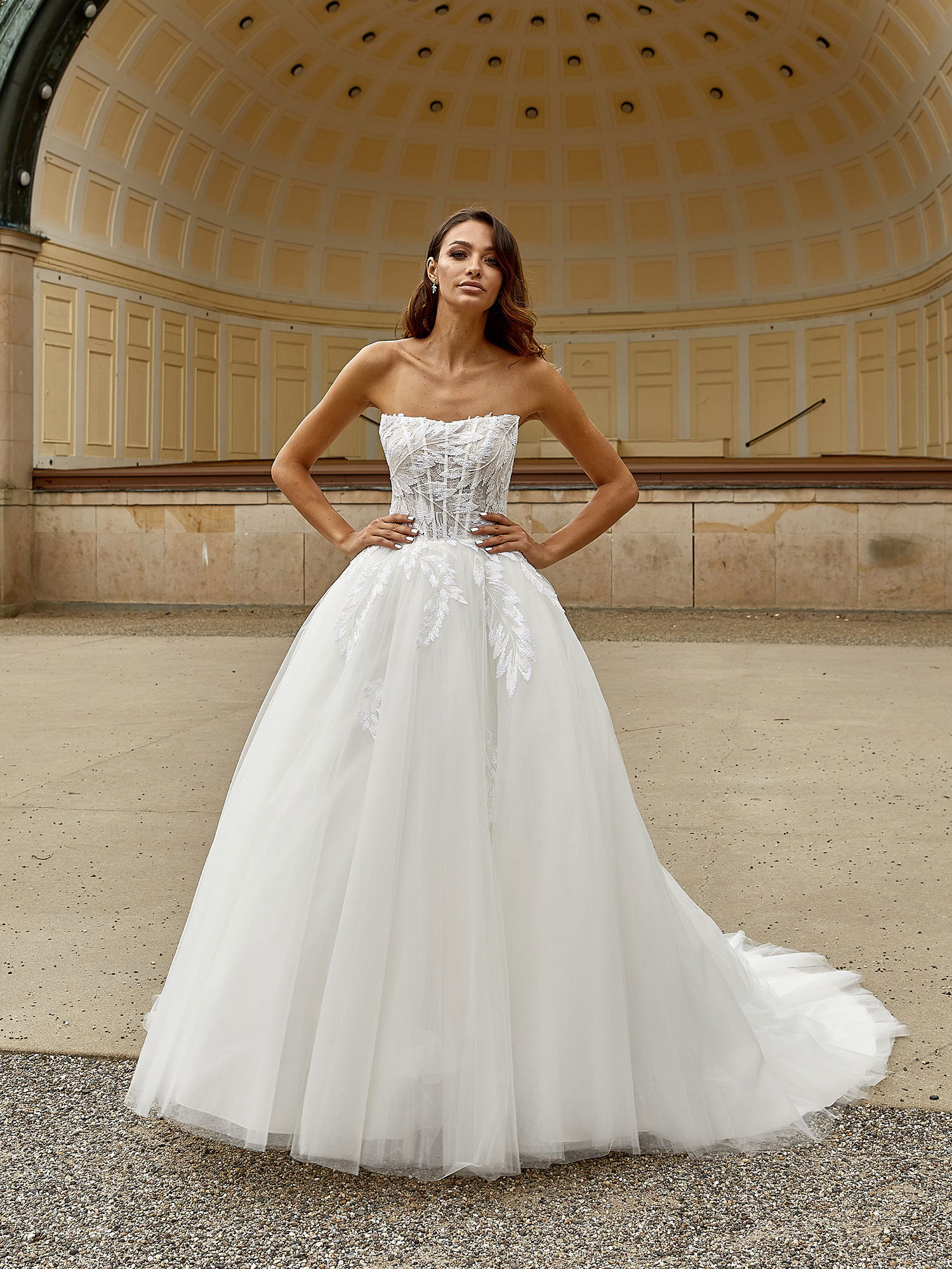 Eine Frau trägt ein trägerloses, korsettartiges Brautkleid mit Spitzenapplikationen und einem weiten Tüllrock. Sie steht vor einem architektonisch auffälligen Pavillon und hat die Hände selbstbewusst auf die Hüften gelegt.