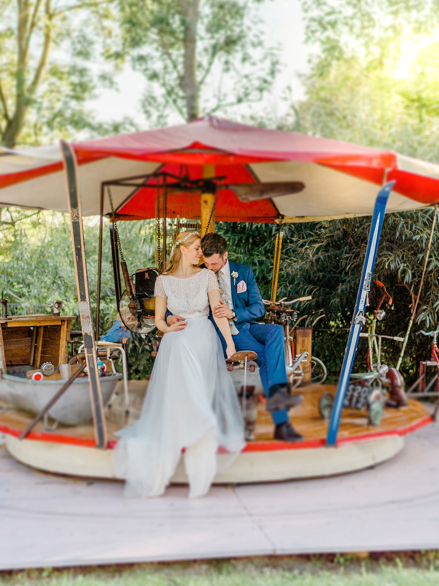 Das Brautpaar sitzt auf einem alten Karussell im Freien. Die Braut in ihrem fließenden Kleid sitzt entspannt, während der Bräutigam sie sanft umarmt. Das Karussell mit rot-weißem Dach verleiht der Szene eine verspielte, nostalgische Atmosphäre. Umgeben von Bäumen wirkt die Szenerie verträumt.