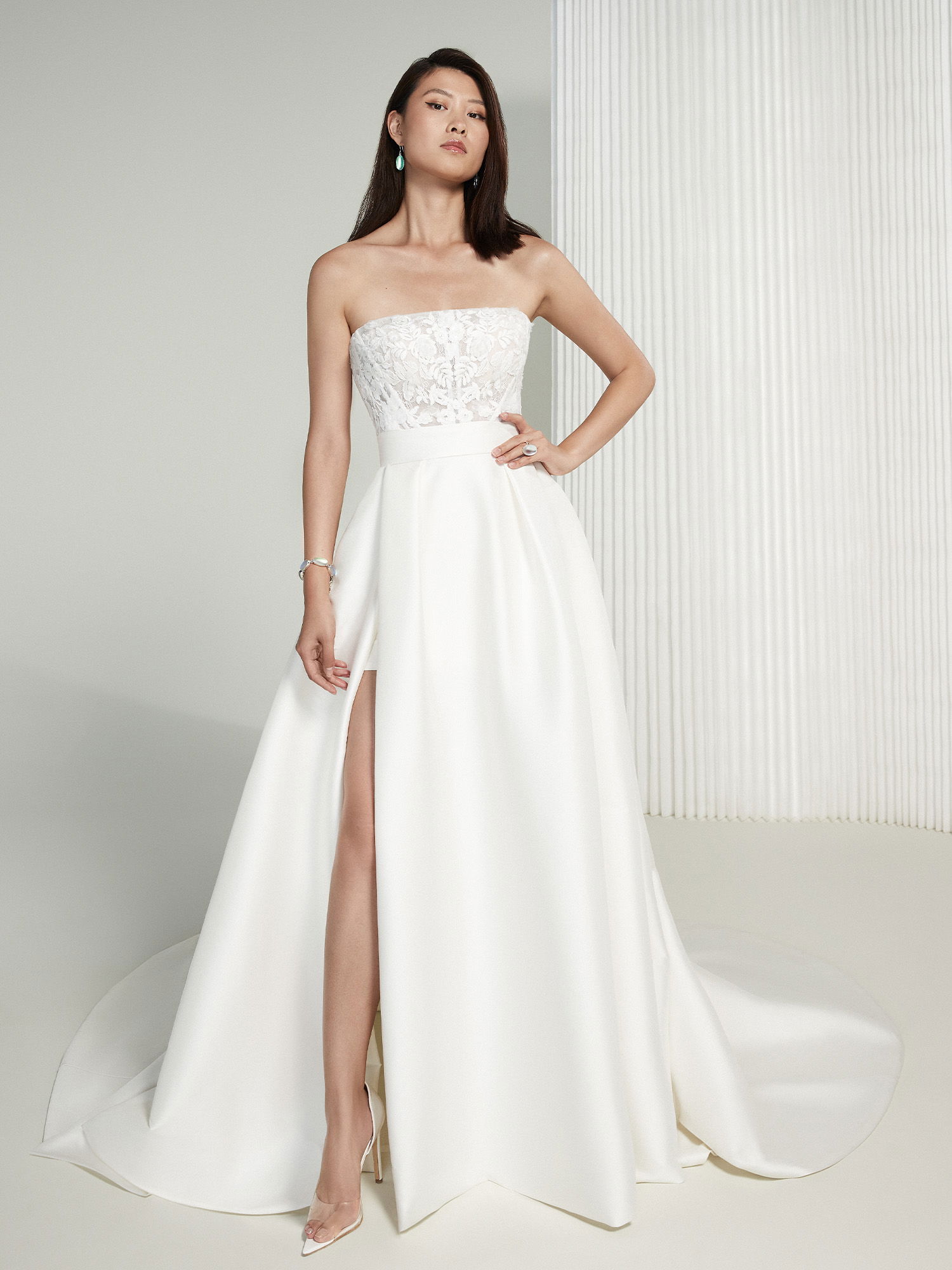 Die Braut trägt ein schulterfreies Kleid mit einer Spitzenkorsage und einem markanten Beinschlitz, der die schlanke Silhouette und die minimalistischen Linien des Outfits unterstreicht.