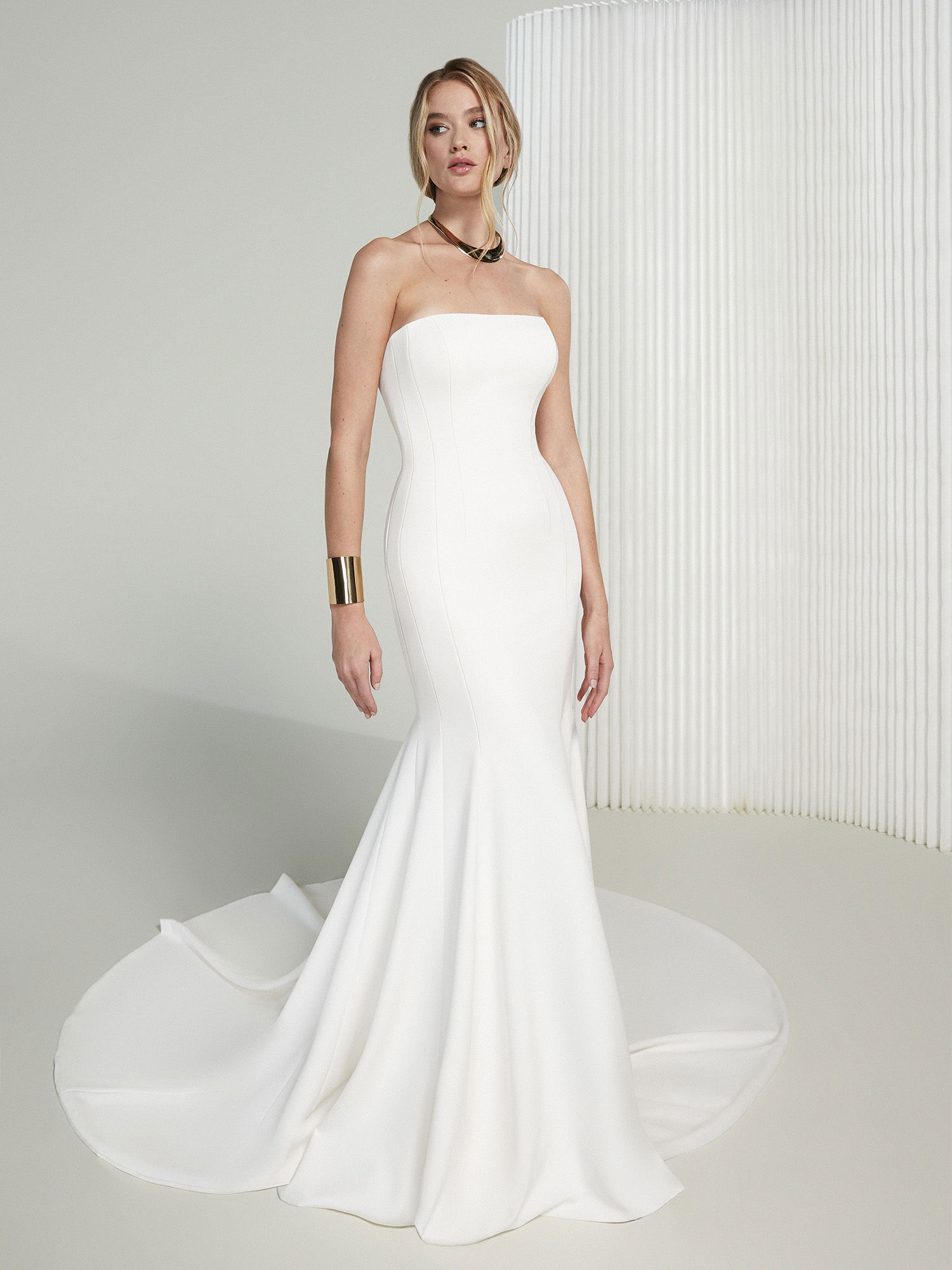 Die Braut posiert in einem modernen, trägerlosen Hochzeitskleid mit Meerjungfrauen-Silhouette. Der einfache Schnitt und die klare Linienführung des Kleides schaffen einen eleganten, zeitlosen Look. Sie kombiniert das Outfit mit auffälligem Goldschmuck, der das Gesamterscheinungsbild betont.