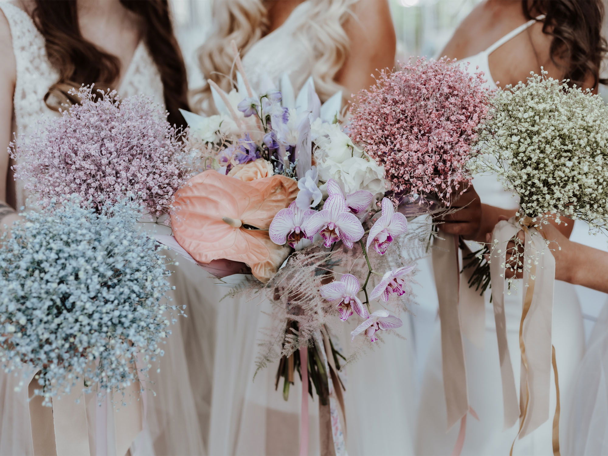 Verschiedene Brautsträuße aus Schleierkraut in Pastellfarben sowie exotische Orchideen und tropische Blätter in den Händen der Bräute.