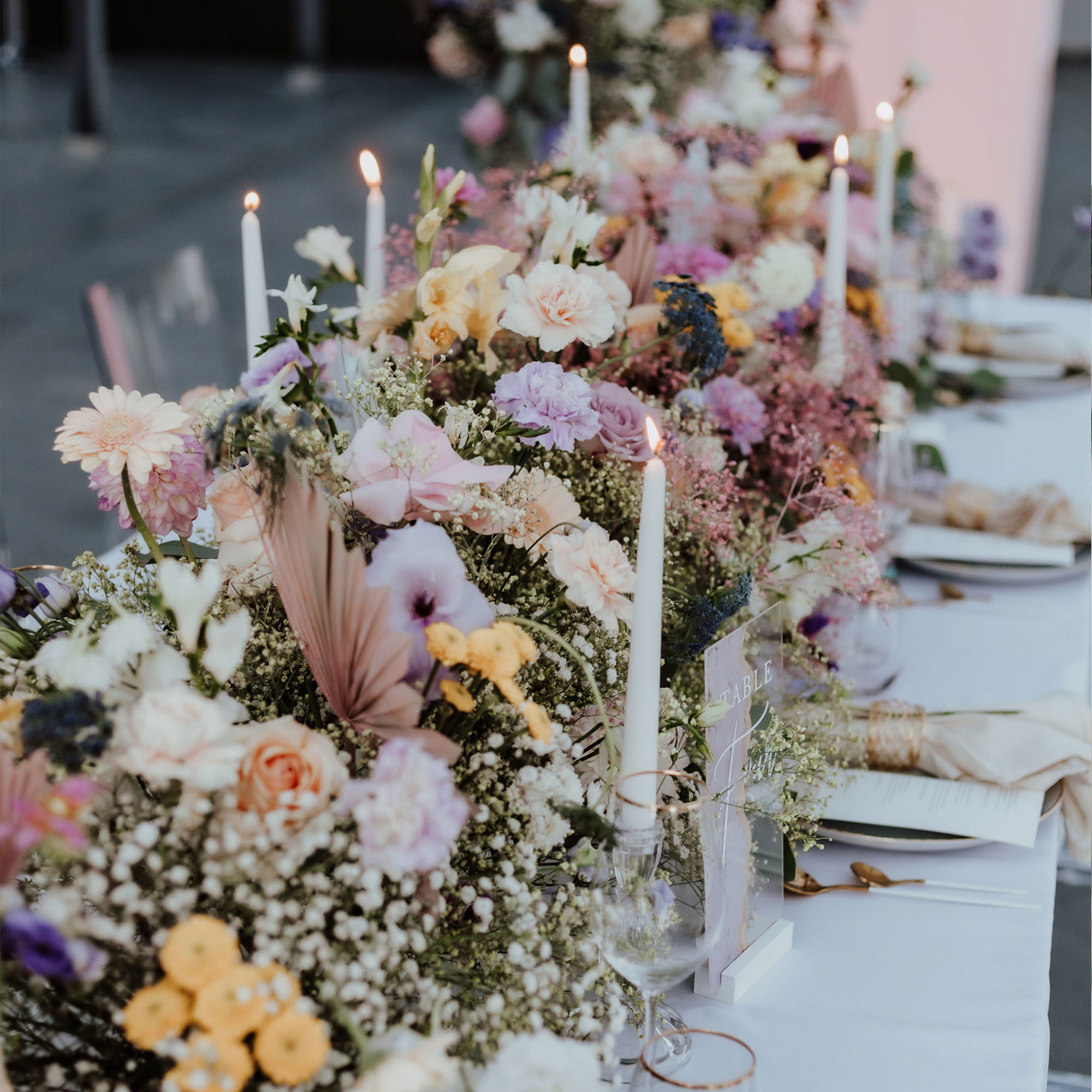 Elegante Hochzeitstischdekoration mit farbenfrohen Blumen in zarten Rosa-, Lila- und Gelbtönen, umrahmt von weißen Kerzen auf einer langen Tafel.