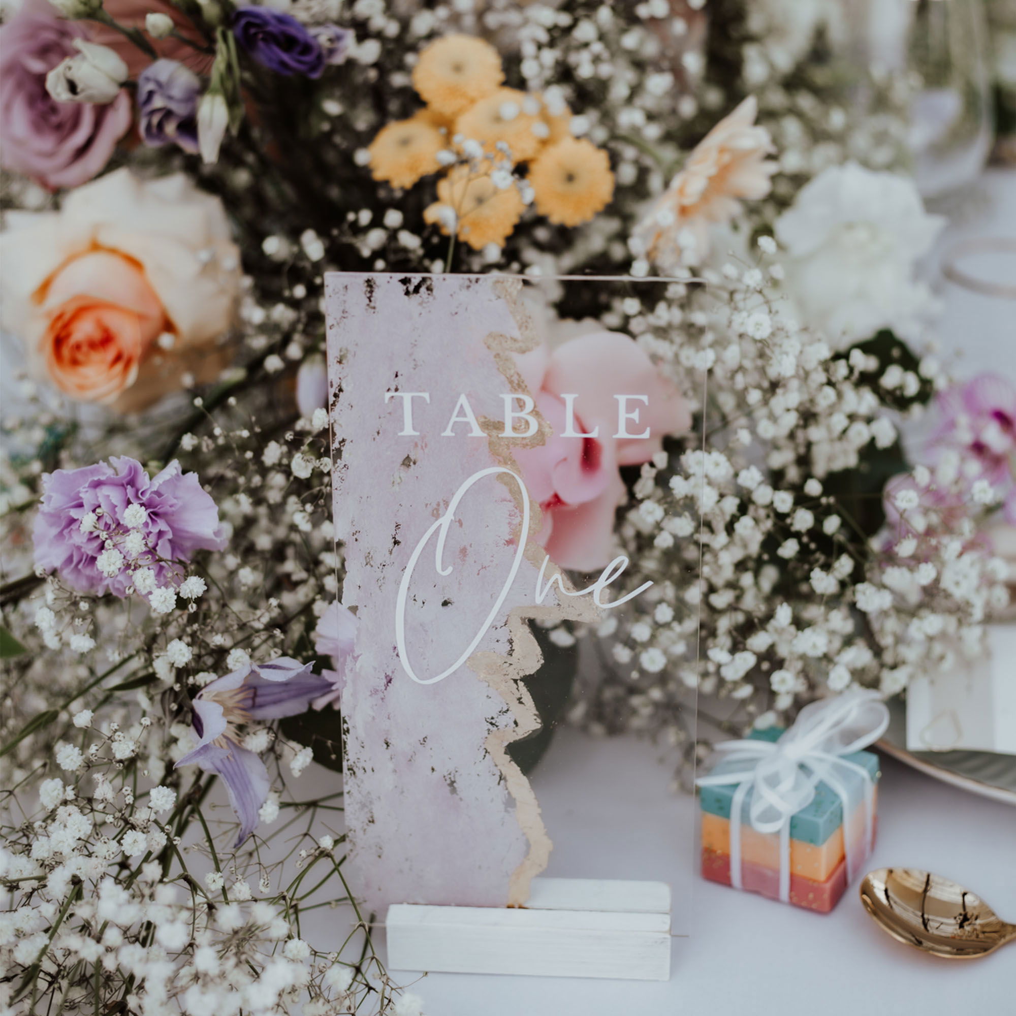 Festliche Tischdekoration mit üppigen Blumenarrangements in sanften Pastelltönen und einer transparenten Tafel mit der Aufschrift 'Table One'.