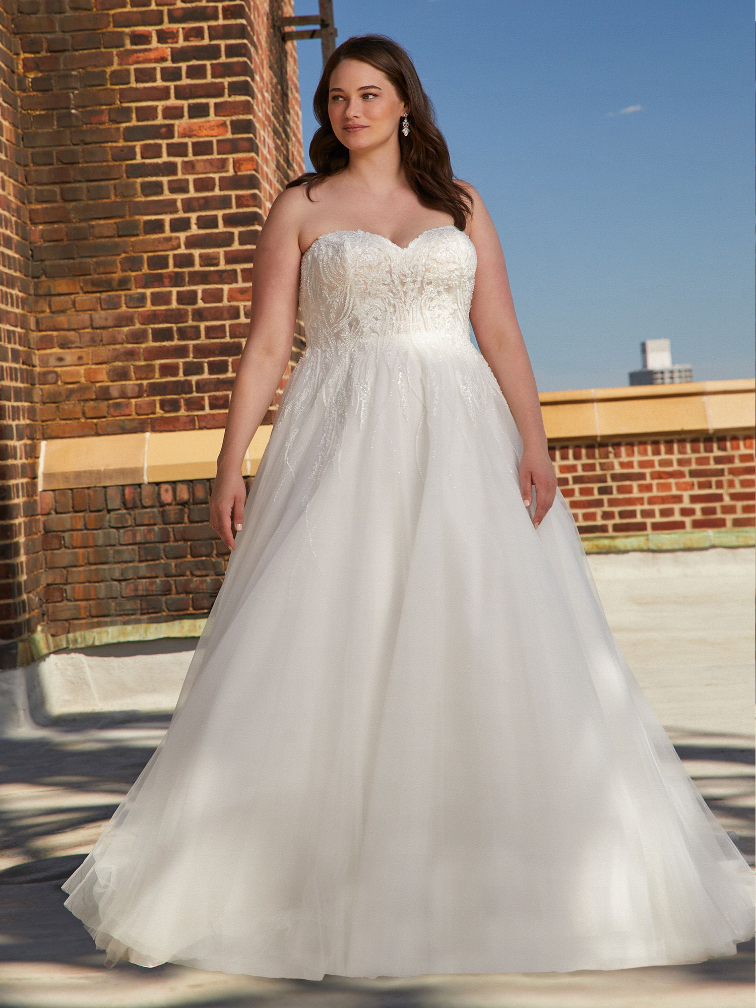 Die Braut präsentiert ein romantisches, trägerloses Brautkleid aus Tüll mit einem detaillierten Oberteil. Im Hintergrund ist eine Backsteinarchitektur zu sehen, die den klassischen Stil des Kleides betont und die urbane Atmosphäre unterstreicht.