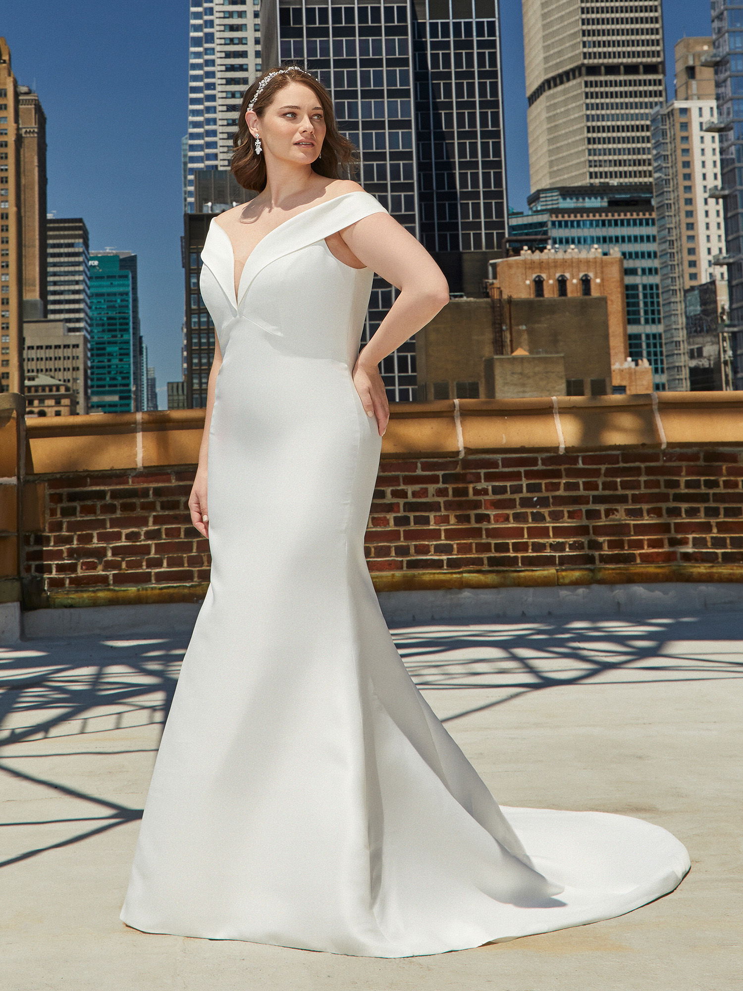 Die Braut präsentiert ein fließendes, modernes Kleid mit offener Schulterpartie und leicht ausgestelltem Rock. Die Szene spielt sich auf einer Terrasse inmitten von Wolkenkratzern ab, die den urbanen Charakter der Umgebung betonen.