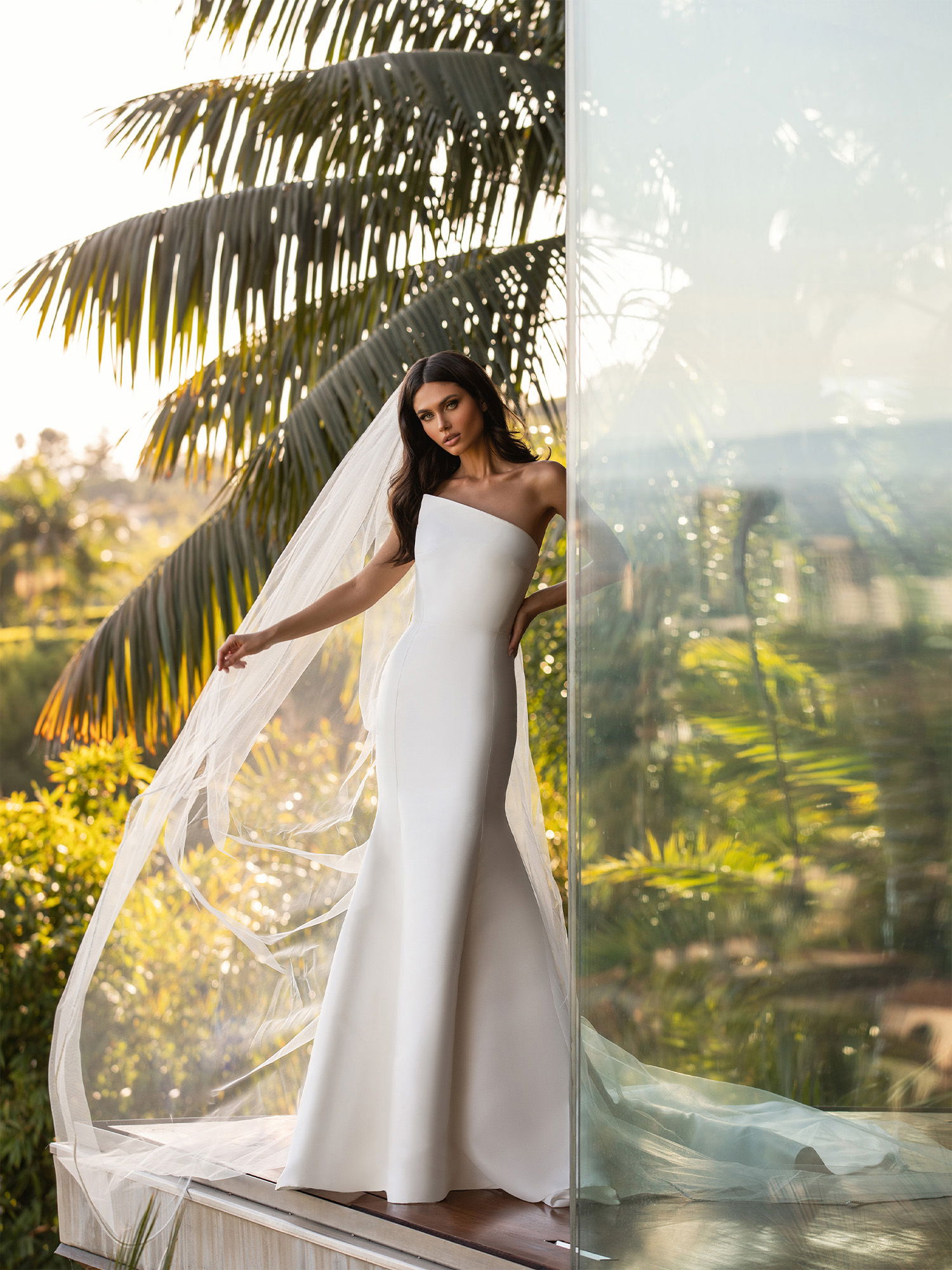 Eine Frau trägt ein minimalistisches, weißes Brautkleid mit asymmetrischer Schulterpartie. Sie posiert neben einer gläsernen Wand, umgeben von tropischen Pflanzen und warmem Sonnenlicht, das die Szene erhellt.