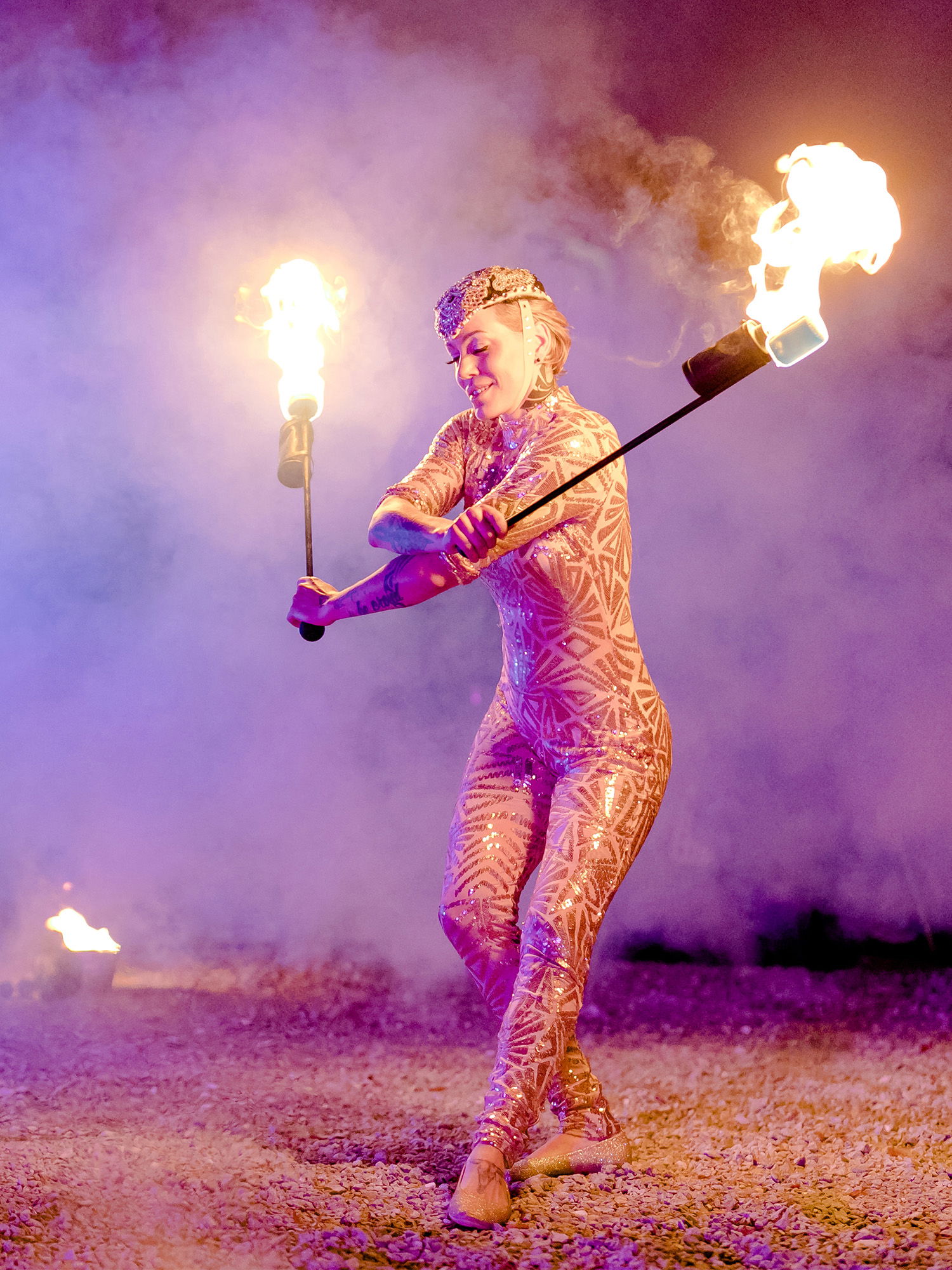 Die Künstlerin, gekleidet in einen schimmernden Overall mit auffälligen Mustern, zeigt eine Feuershow, bei der sie zwei brennende Stäbe kunstvoll um sich schwingt. Rauch steigt in die Luft und das warme Licht der Flammen schafft eine mystische und fesselnde Szenerie.