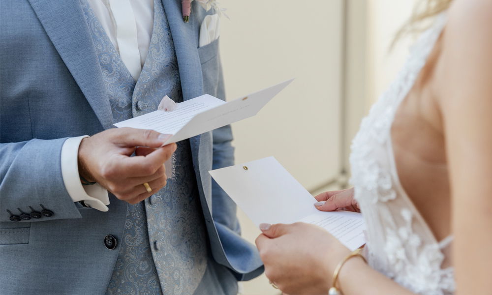 Braut und Bräutigam im eleganten Hochzeitsoutfit halten ihre persönlichen Eheversprechen während der Zeremonie in den Händen.