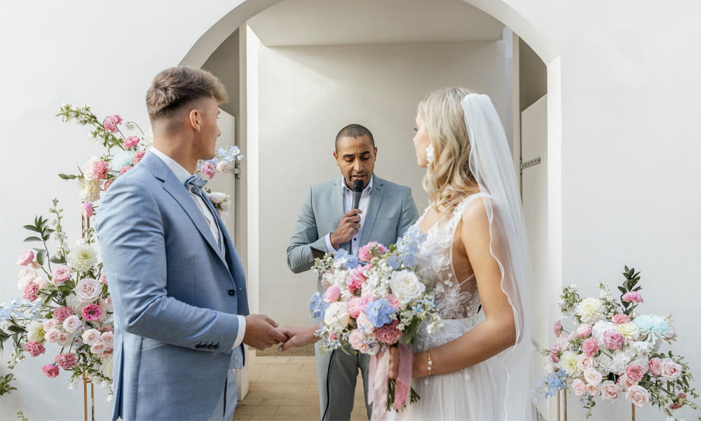 Brautpaar steht während der Trauung vor dem Trauredner, umgeben von üppiger Blumendekoration in Pastelltönen.