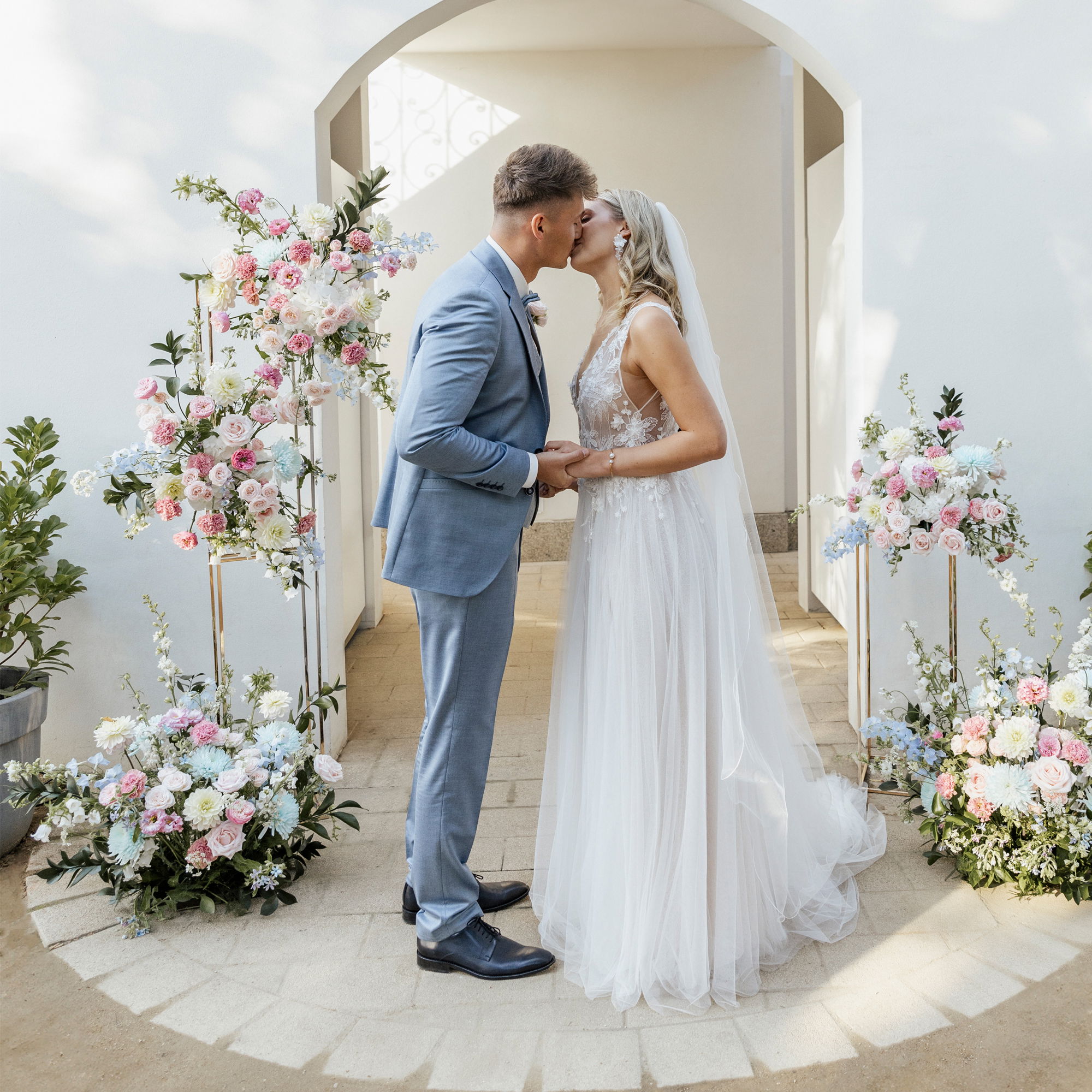 Braut und Bräutigam küssen sich bei der Trauung unter einem weißen Bogen, umgeben von üppiger Blumendeko in Pastelltönen.