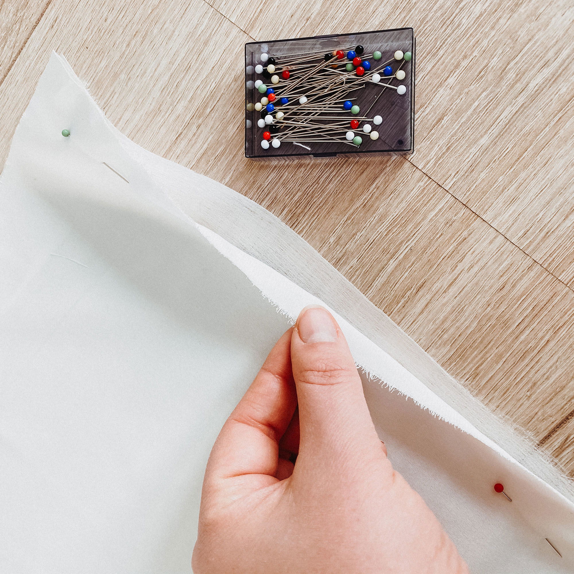 Nahaufnahme einer Hand, die einen weißen Stoff hält, während Stecknadeln für das Nähen bereitliegen. Der Stoff wird über einem Holzboden ausgebreitet.