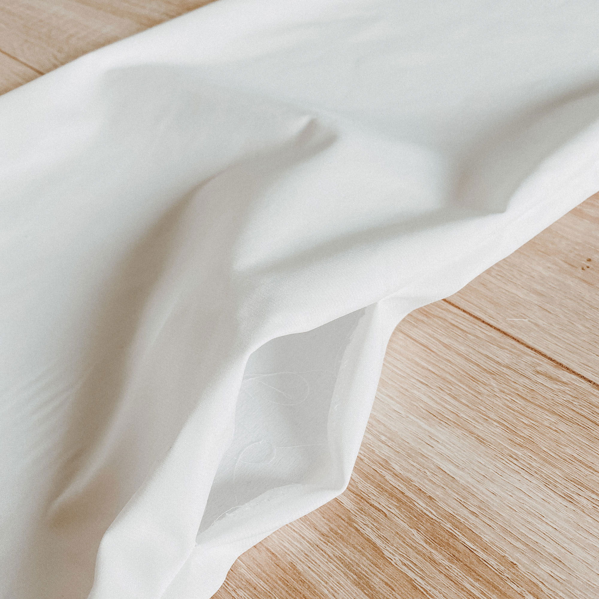 Glatter, weißer Stoff ist locker auf einem hellen Holzboden drapiert, wobei die sanften Wellen des Materials hervorgehoben werden.