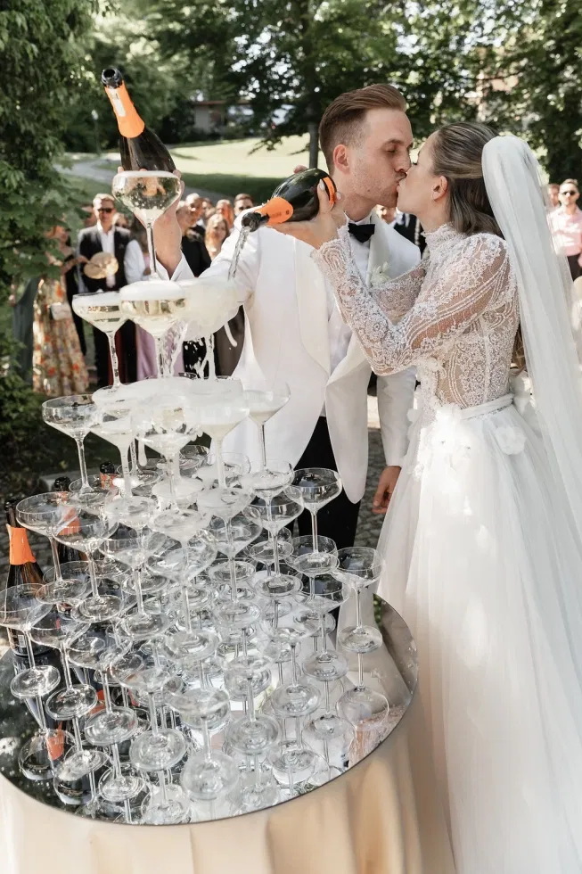 Braut und Bräutigam küssen sich beim Einschenken von Champagner in eine große Glaspyramide vor ihren Hochzeitsgästen im Garten.
