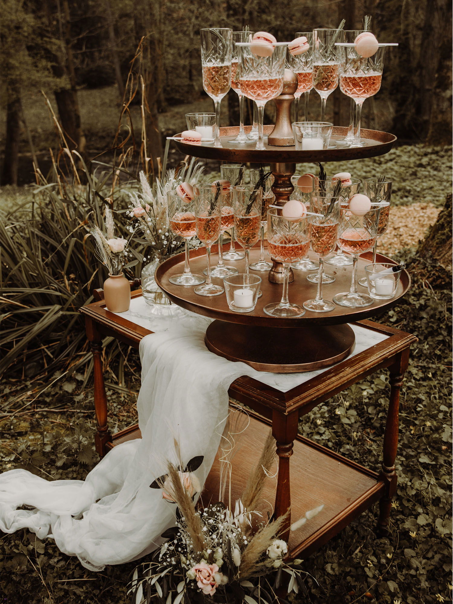 Champagner-Turm mit Kristallgläsern, gefüllt mit roséfarbenem Sekt, auf einem rustikalen Holztisch, umrahmt von einem natürlichen Waldhintergrund. Die Gläser sind liebevoll mit Macarons dekoriert, und die Szene ist mit weißen Leinentüchern und Blumen geschmückt.