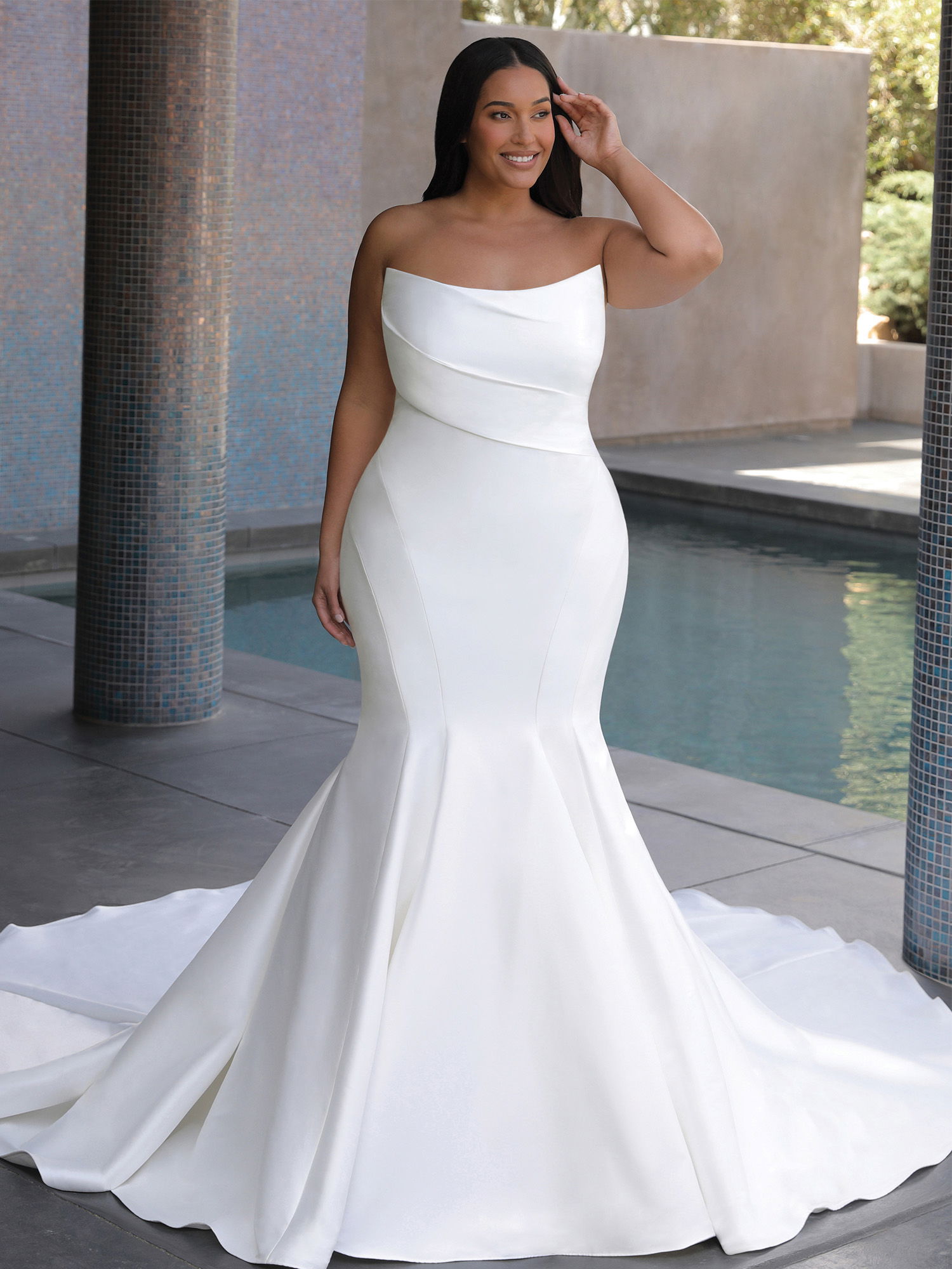 Eine Frau in einem eleganten, trägerlosen Brautkleid mit sanft drapiertem Oberteil steht auf einer modernen Terrasse. Im Hintergrund sind Fliesen und eine moderne Architektur sichtbar, während das Kleid stilvoll auf dem Boden fällt.