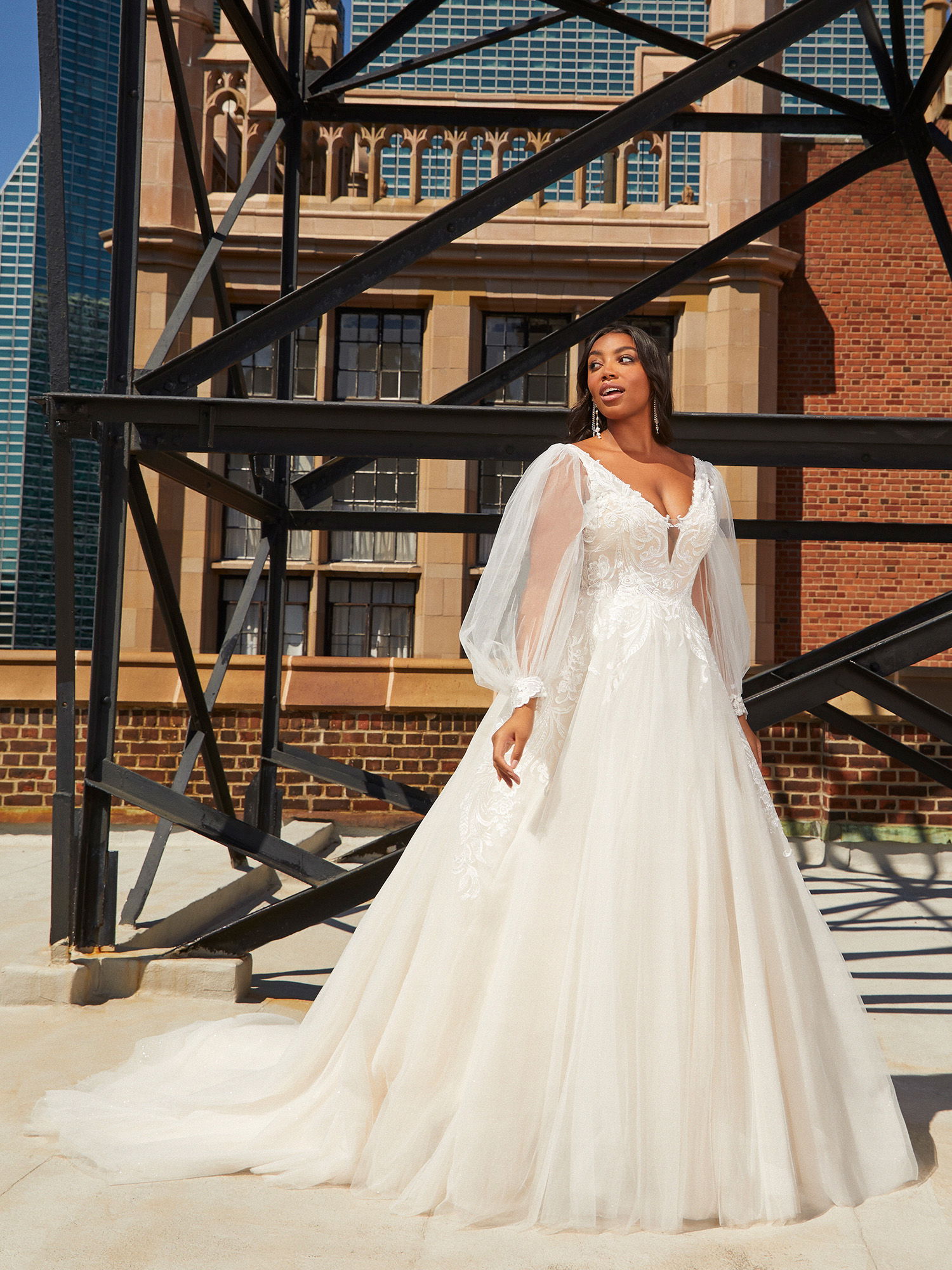 Die Braut trägt ein atemberaubendes Kleid mit transparenten Ärmeln und einer weiten Schleppe. Sie steht vor einem Gebäude mit Ziegelwänden und Metallstrukturen, die das Licht reflektieren und die Eleganz des Moments hervorheben.