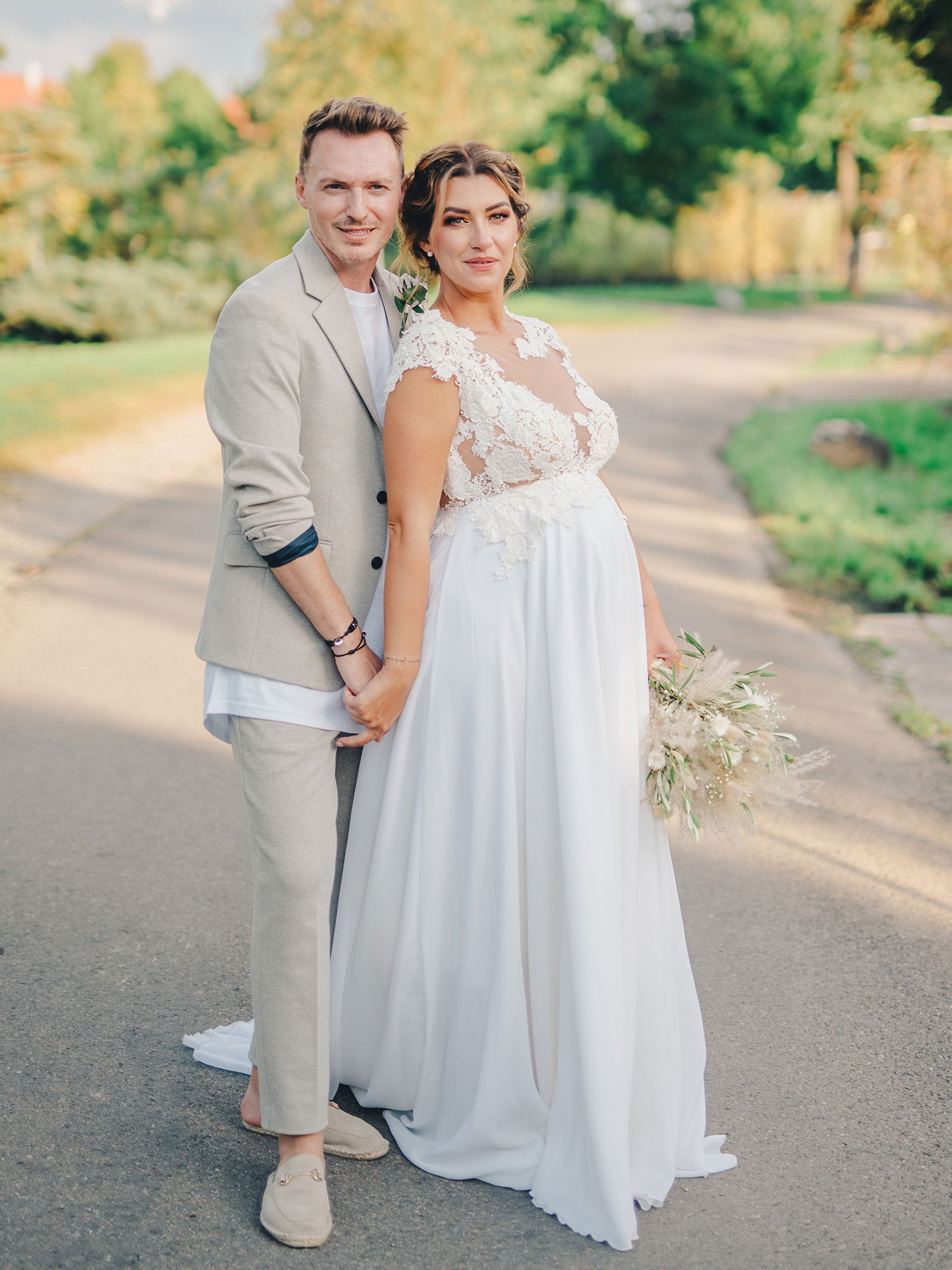 Ein lächelndes Hochzeitspaar posiert auf einem Landweg, die Braut trägt ein romantisches weißes Kleid mit einem Spitzenoberteil, während der Bräutigam in einem lässigen, hellen Anzug gekleidet ist. Beide halten sich an den Händen.