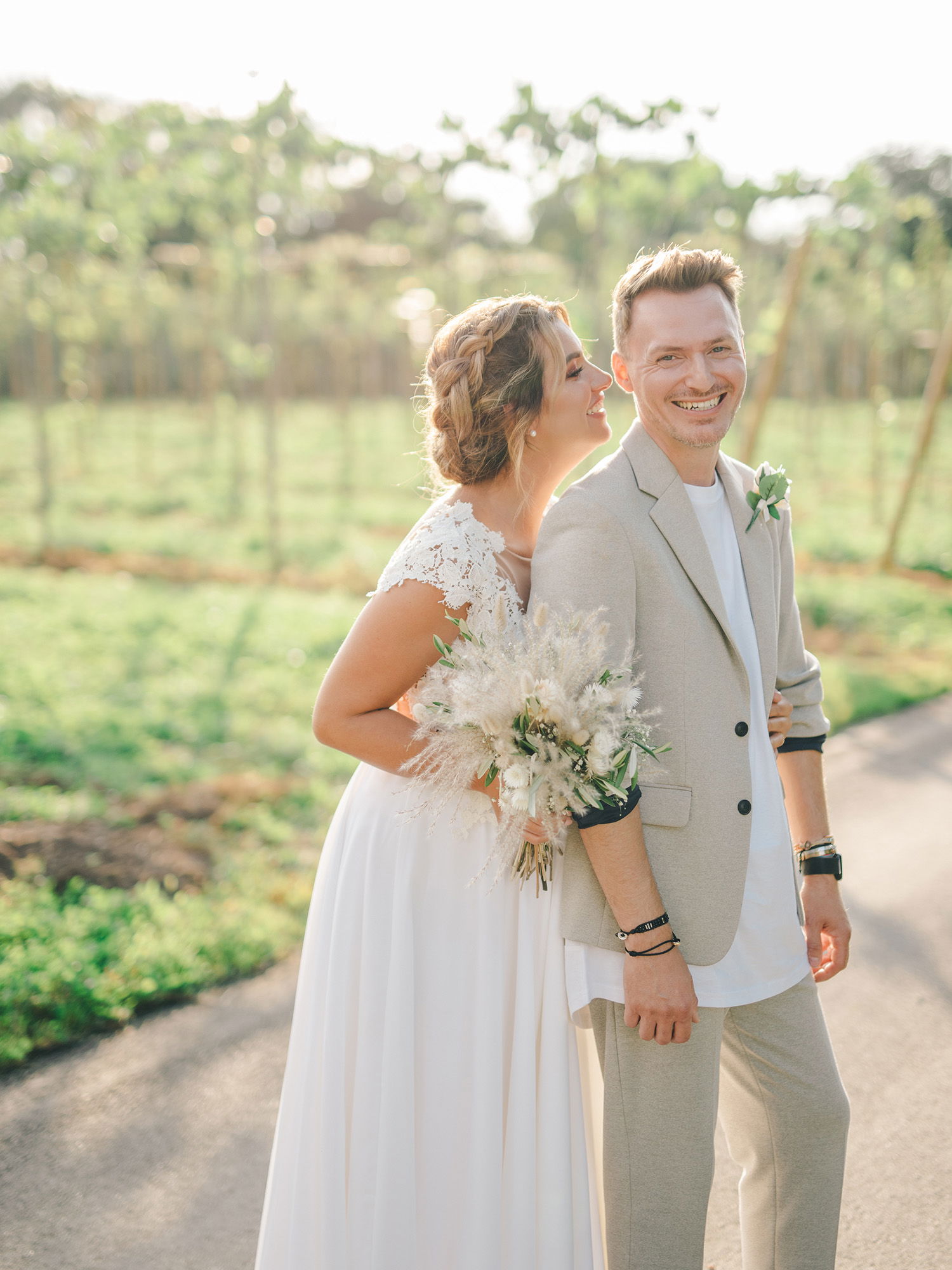 Ein glückliches Brautpaar posiert in der Natur, umgeben von grünen Feldern. Die Braut hält einen Strauß aus Trockenblumen und lächelt, während sie sich an den Bräutigam lehnt, der in einem lässigen, hellen Anzug gekleidet ist.