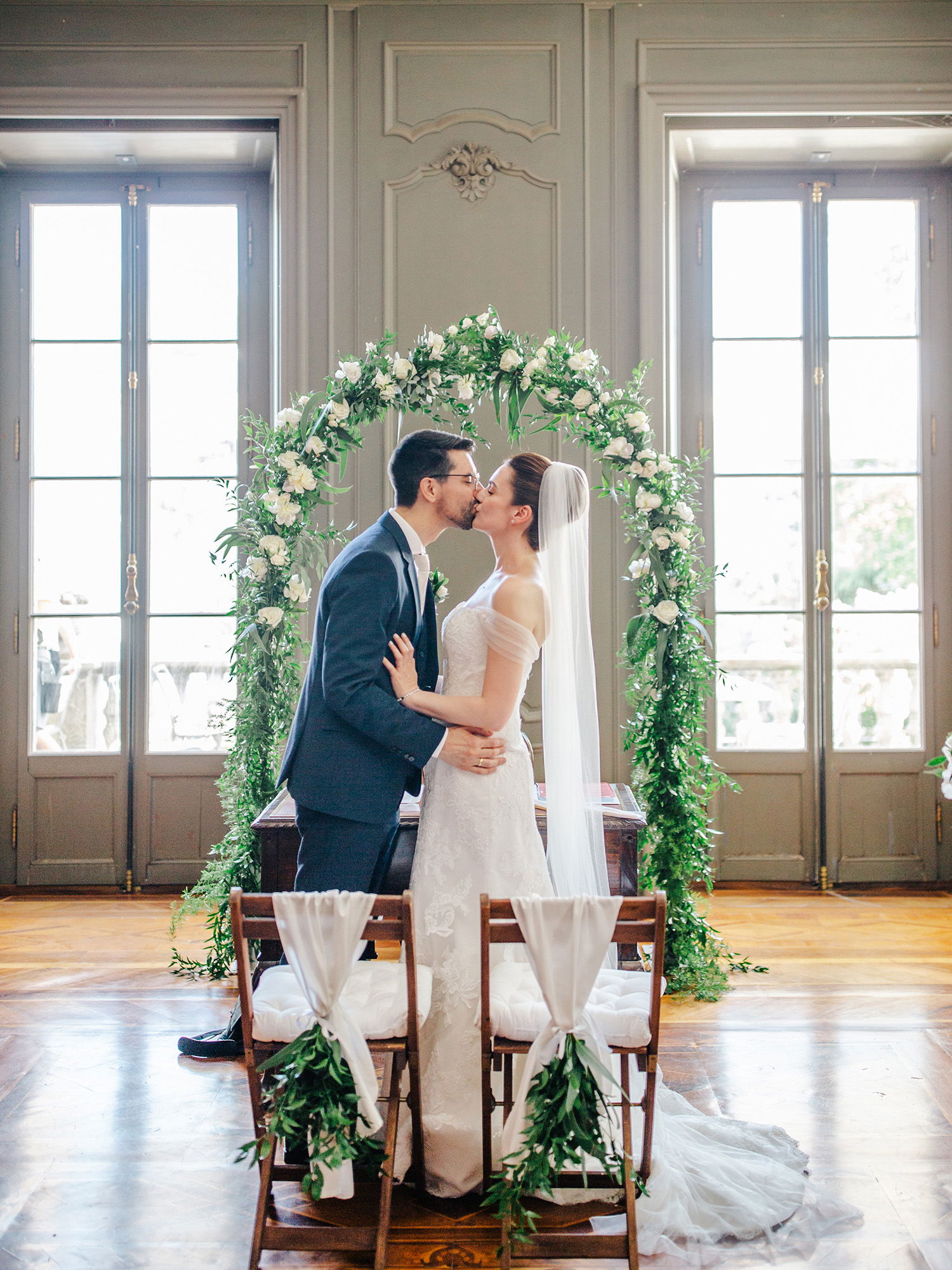 Ein verliebtes Paar tauscht einen Kuss vor einem üppigen Blumenbogen bei der Hochzeit. Die Braut in einem weißen Kleid und Schleier steht mit dem Bräutigam in einem eleganten Anzug in einem lichtdurchfluteten Raum mit hohen Fenstern.