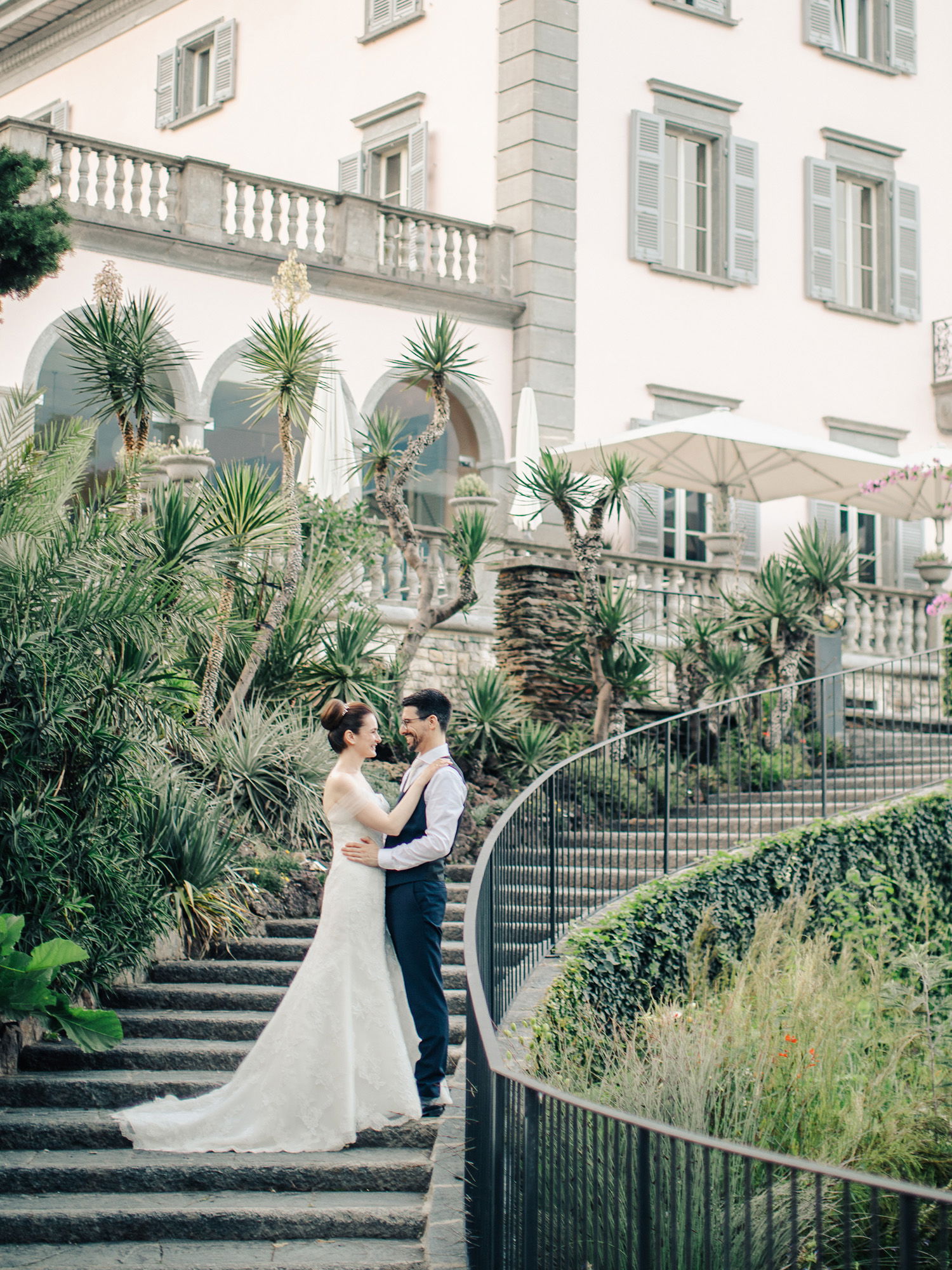 Ein Hochzeitspaar posiert auf einer geschwungenen Steintreppe vor einer historischen Villa. Die Braut in einem eleganten Spitzenkleid schaut den Bräutigam an, der einen blauen Anzug trägt. Tropische Pflanzen säumen die Szene.