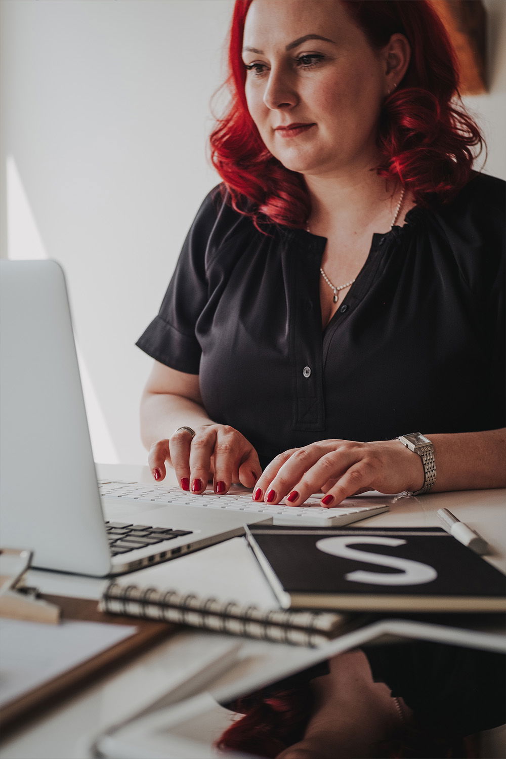 Eine Frau mit leuchtend roten Haaren und einer schwarzen Bluse tippt konzentriert auf ihrem Laptop. Auf dem Schreibtisch vor ihr liegen Notizbücher und ein Stift.