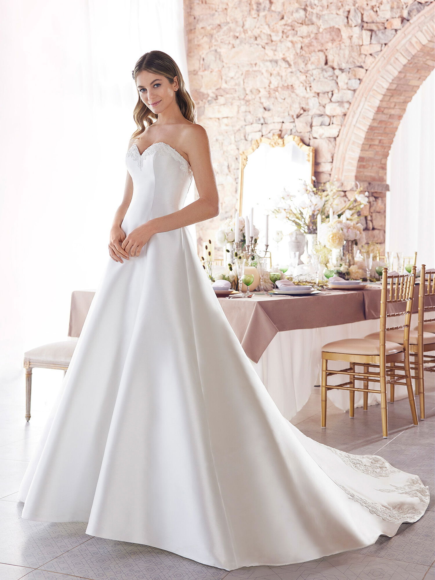 Eine Braut in einem schlichten, eleganten A-Linien-Kleid mit herzförmigem Ausschnitt posiert in einem hellen Raum. Das Kleid hat eine lange Schleppe, und die Braut steht vor einem rustikalen Tisch mit Blumen und Kerzen.