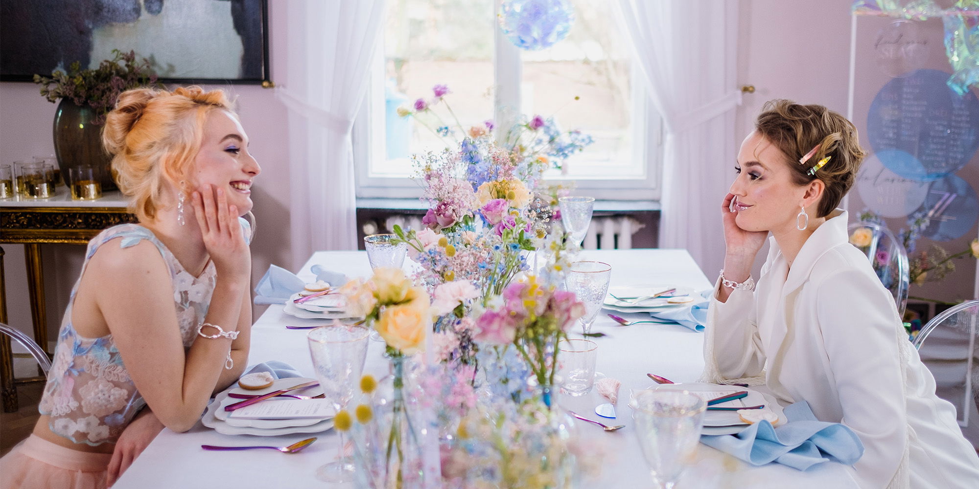 Zwei Freundinnen sitzen an einem hellen, fröhlich dekorierten Tisch voller Blumen in zarten Farben. Sie wirken entspannt und lachen miteinander. Durch das Fenster hinter ihnen strahlt sanftes Tageslicht in den Raum, während bunte Blumen den Tisch in Szene setzen.
