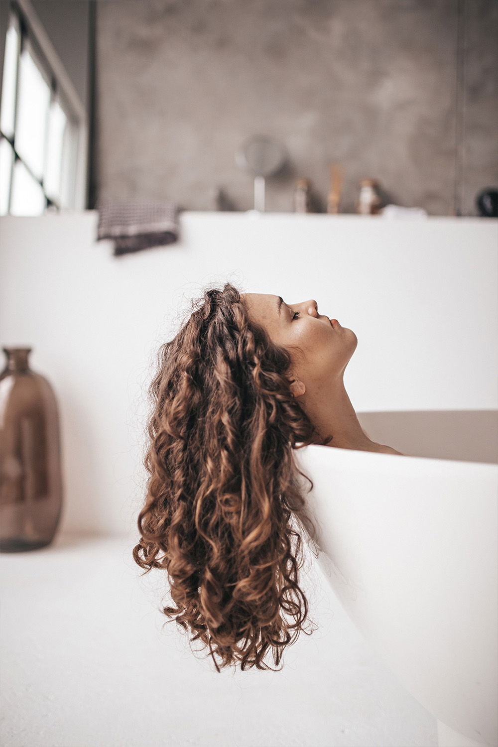 Eine Frau mit langen, lockigen Haaren ruht in einer weißen Badewanne. Ihr Kopf ist nach hinten geneigt, während sie sich entspannt. Die minimalistische Einrichtung und das weiche Licht sorgen für eine ruhige Atmosphäre.