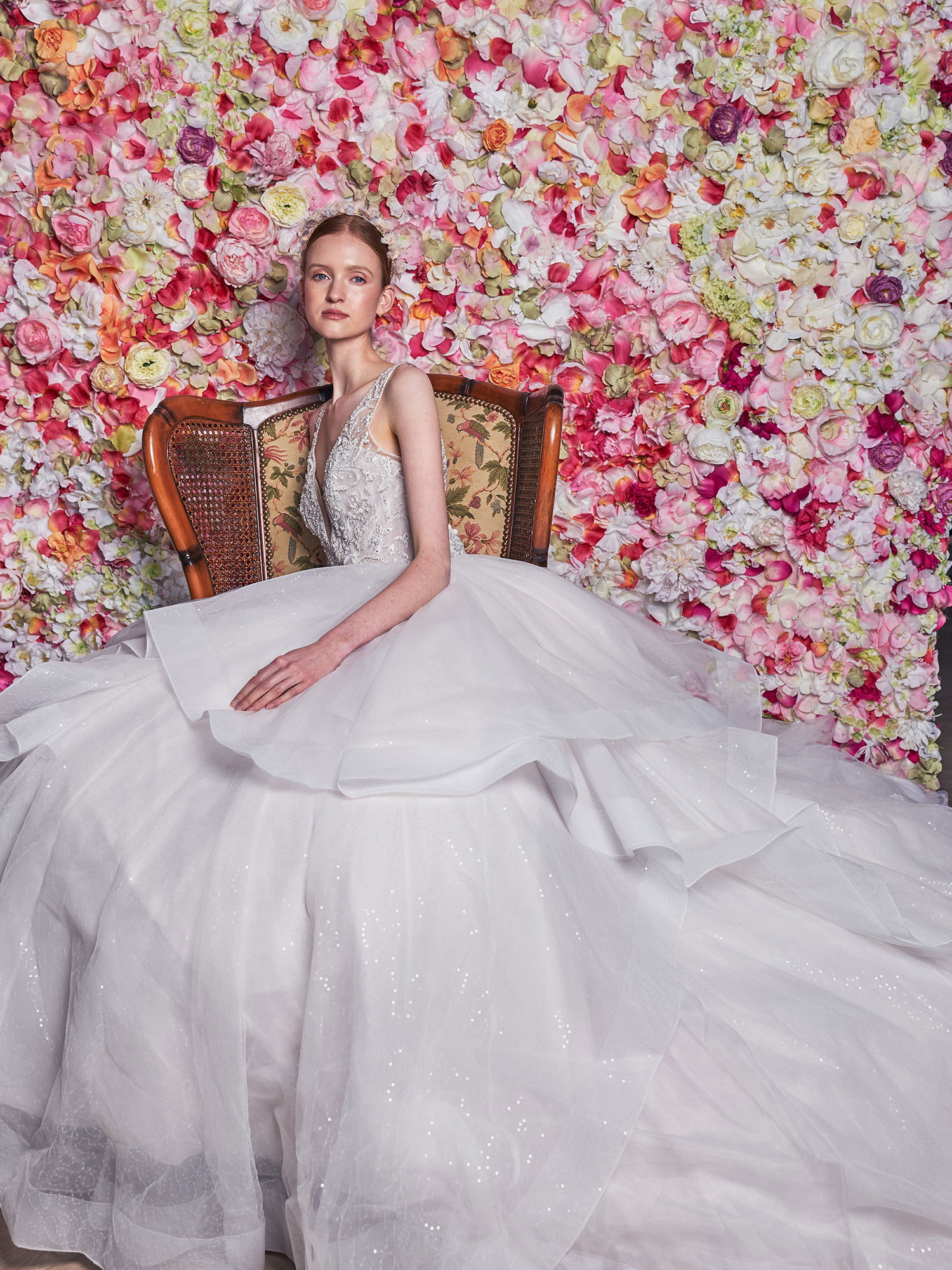 Eine junge Braut mit einem aufwändigen, voluminösen Brautkleid sitzt auf einem Stuhl vor einer Wand, die mit unzähligen Ranunkeln in verschiedenen Farben bedeckt ist. Ihr Kleid ist auslagenreich und elegant, während die Blumenwand im Hintergrund eine romantische Atmosphäre schafft.